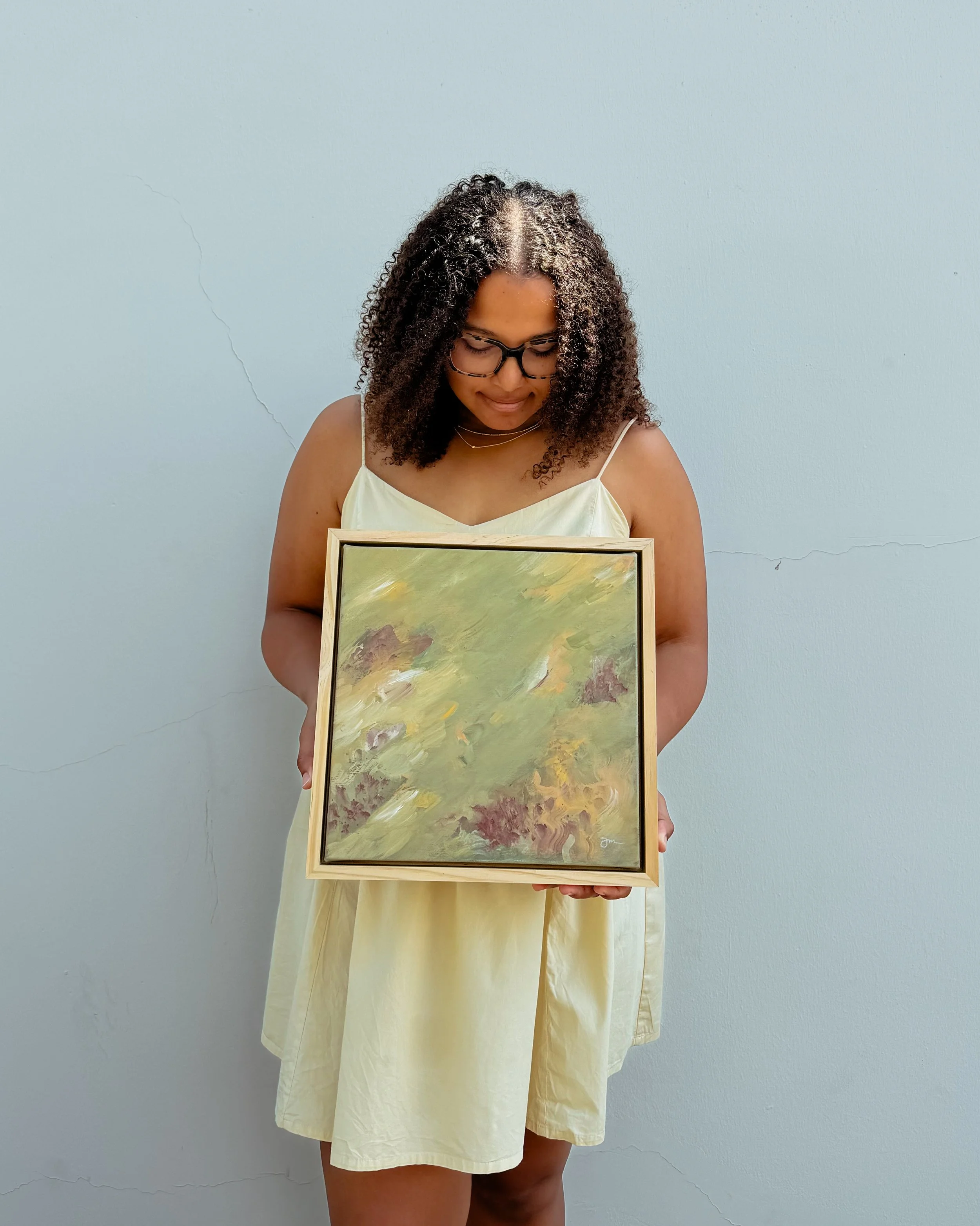 A woman with curly hair, glasses, and wearing a light yellow dress is holding a framed abstract painting with green, yellow, and orange and red colors, standing against a plain wall.