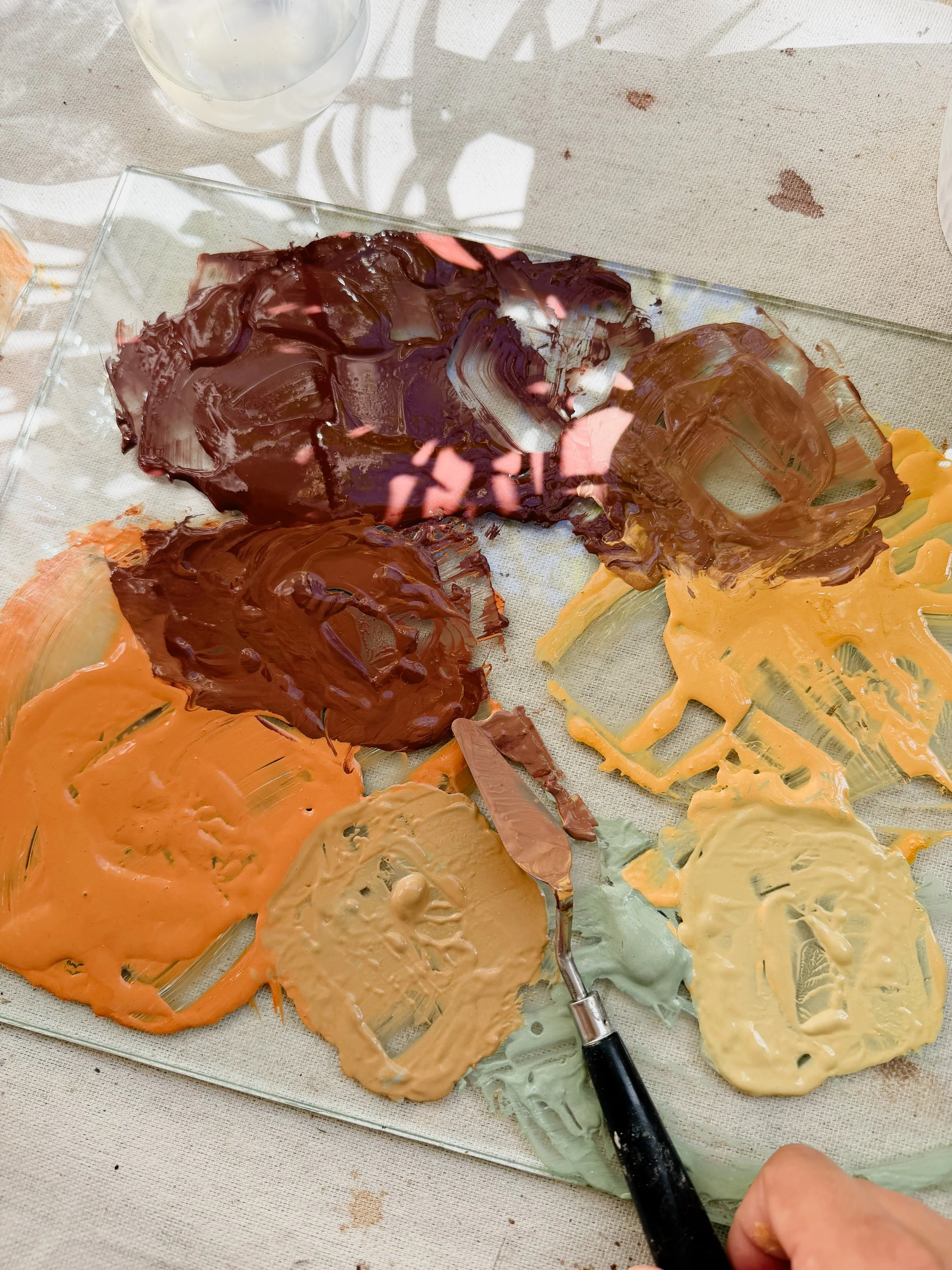 Artist mixing different colored paints on a glass palette using a palette knife, with a hand holding the knife visible in the bottom right corner.