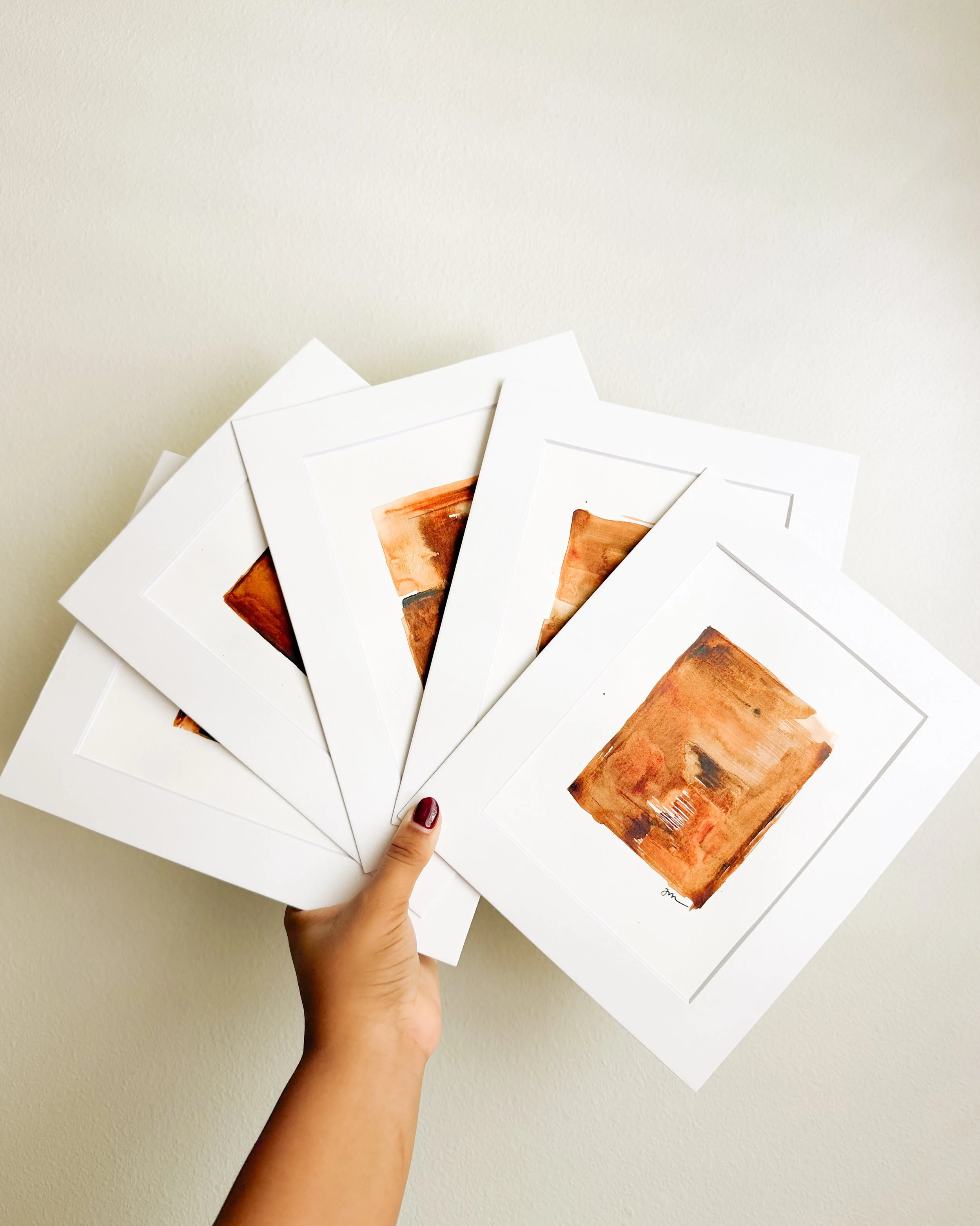 A hand holding several pieces of white paper with brown orange and red color artworks on them against a plain wall background.