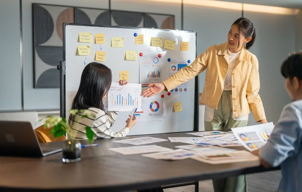 A woman presents data on a whiteboard to two colleagues in a meeting room. The whiteboard has charts, graphs, and sticky notes. The table has documents, a laptop, and a plant.