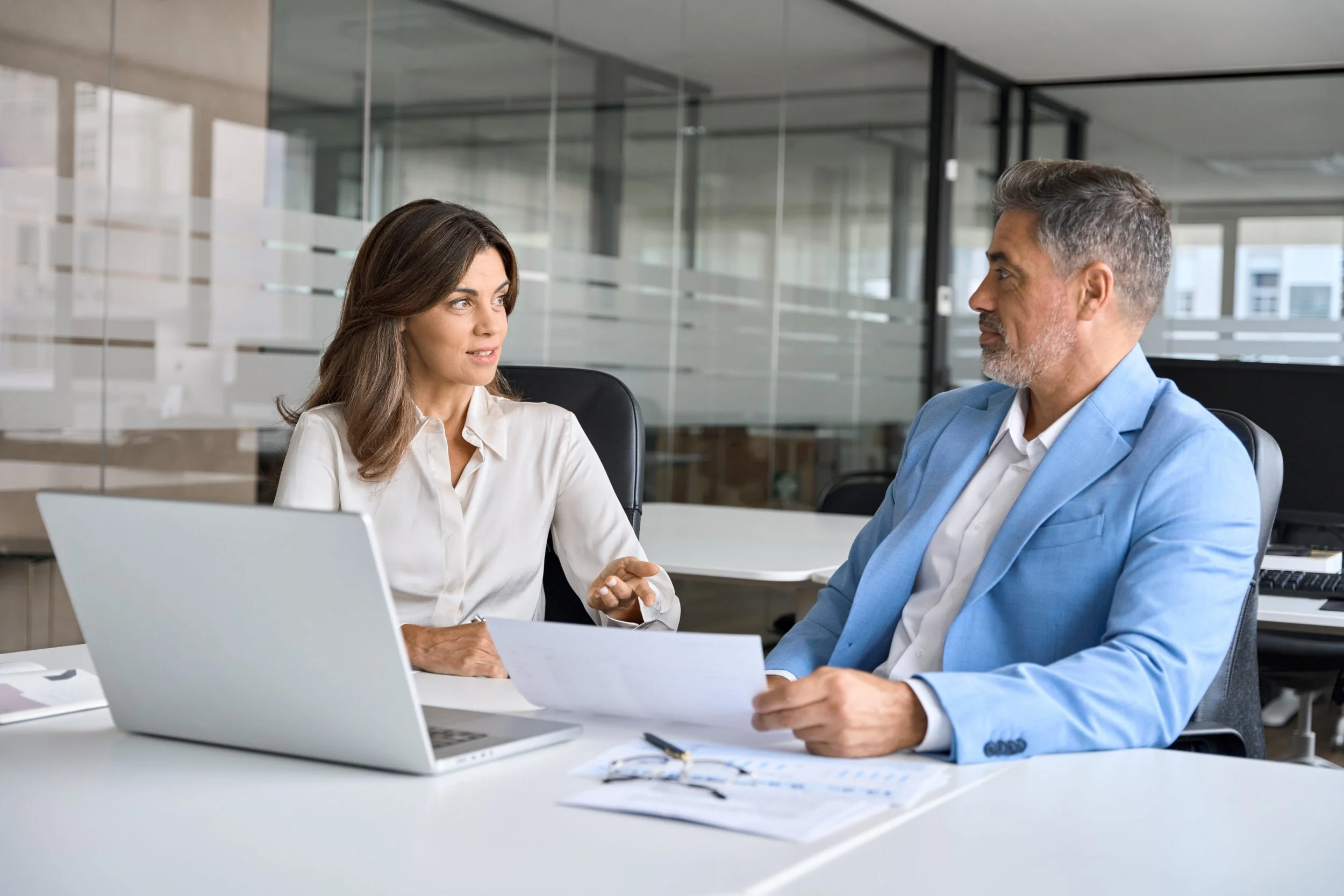 Two professionals, a woman and a man, are seated at a desk in a modern office, engaging in a conversation. The woman has long brown hair and is dressed in a white blouse, while the man has short gray hair and is wearing a light blue blazer over a white shirt. There is a laptop, some documents, and a notepad on the desk.