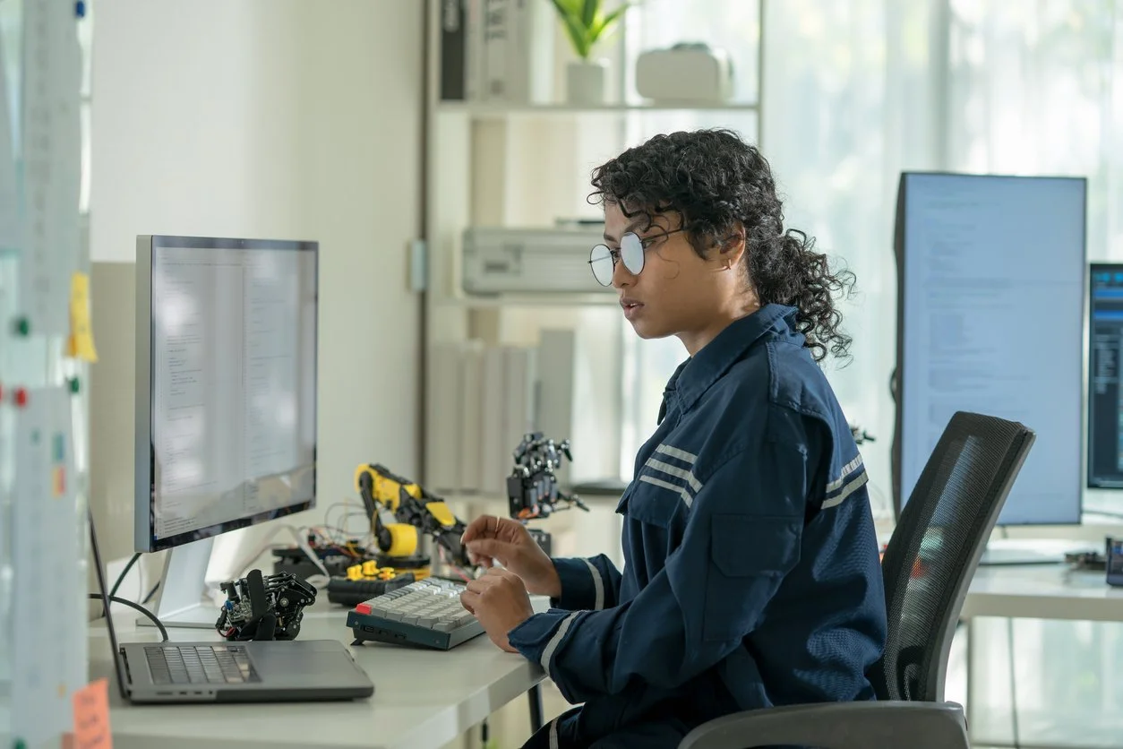 Young woman in a navy blue work uniform sitting at a desk with multiple computer monitors, working on electronic components and repair tools in a well-lit office.