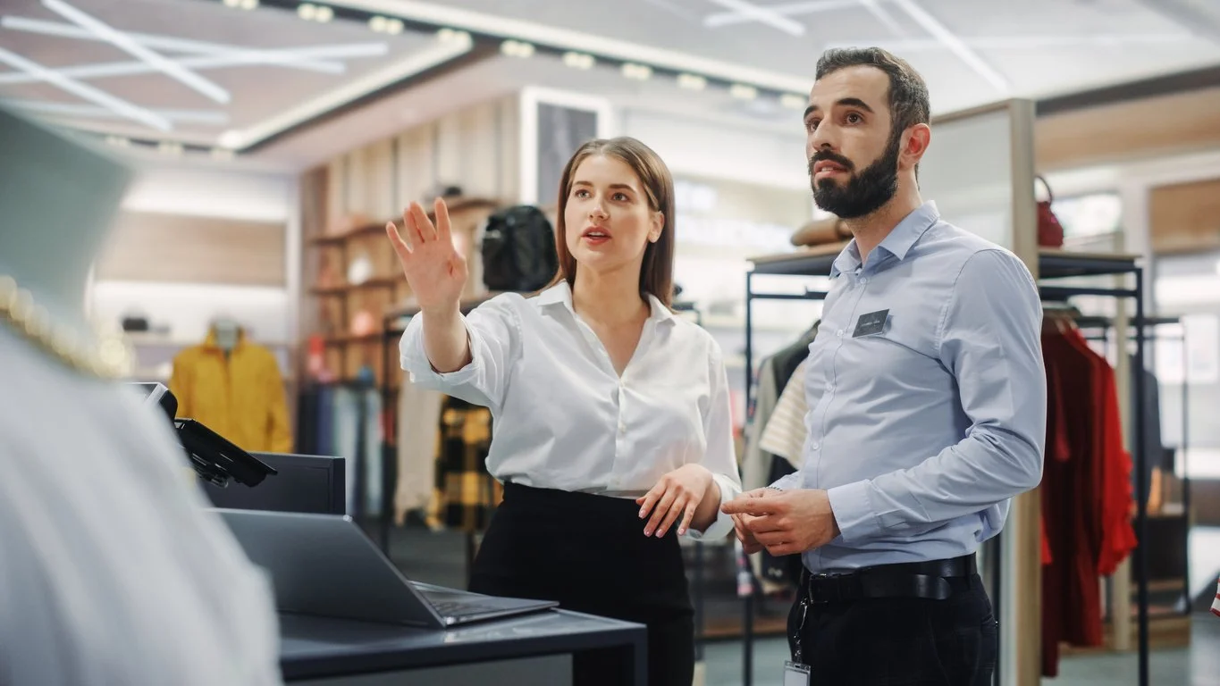 A woman and a man are working at a clothing store counter, with the woman gesturing with her right hand and the man holding a small object in his hands, while discussing or explaining something to a customer.