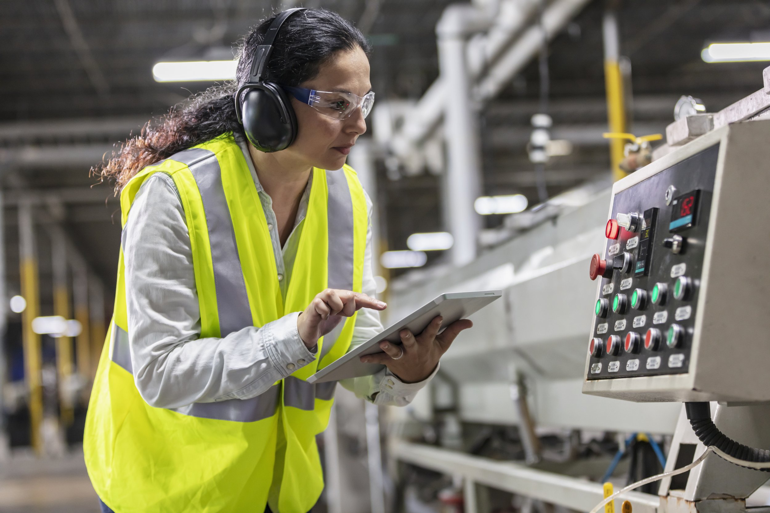 Woman in safety gear inspecting control panel in industrial setting.