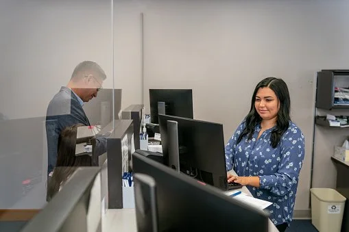 A man and a woman working at computer stations separated by a glass partition in a bank.