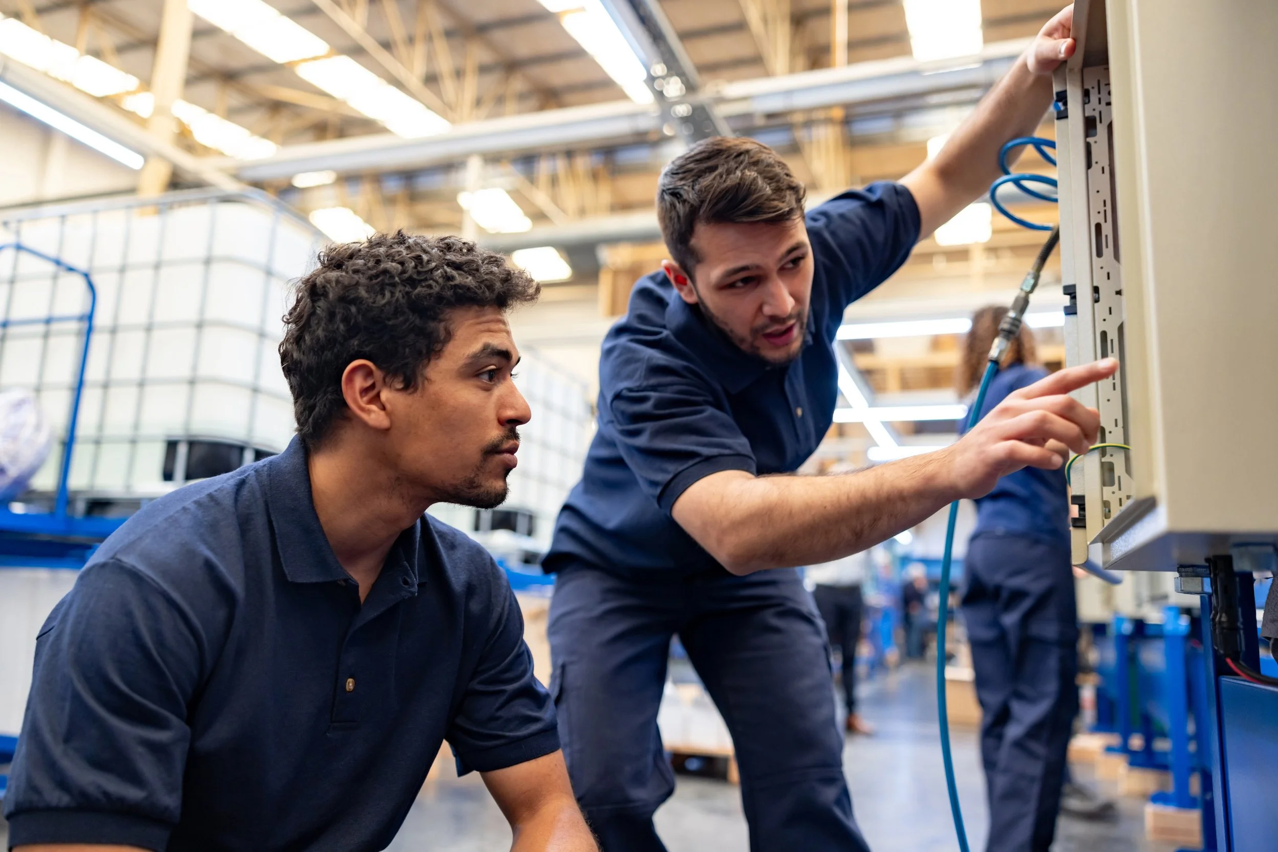 Two men in blue work uniforms are working together on electrical wiring inside an industrial or warehouse environment. One man is closely examining the wires while the other is reaching into an electrical panel.