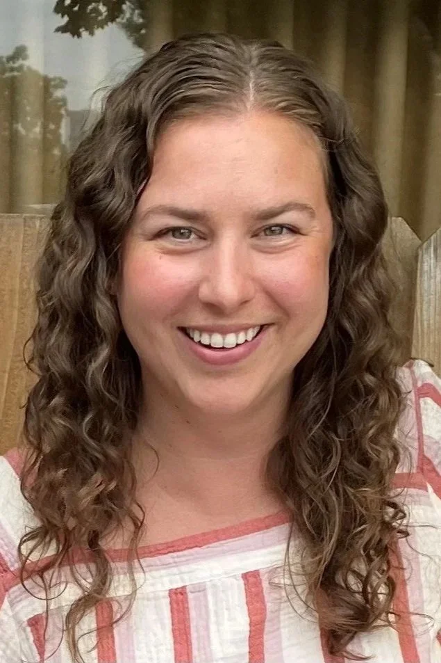 Emily Segel smiling with long, curly brown hair and a broad smile, wearing a white and pink striped top, sitting outdoors with a wooden fence and trees in the background.