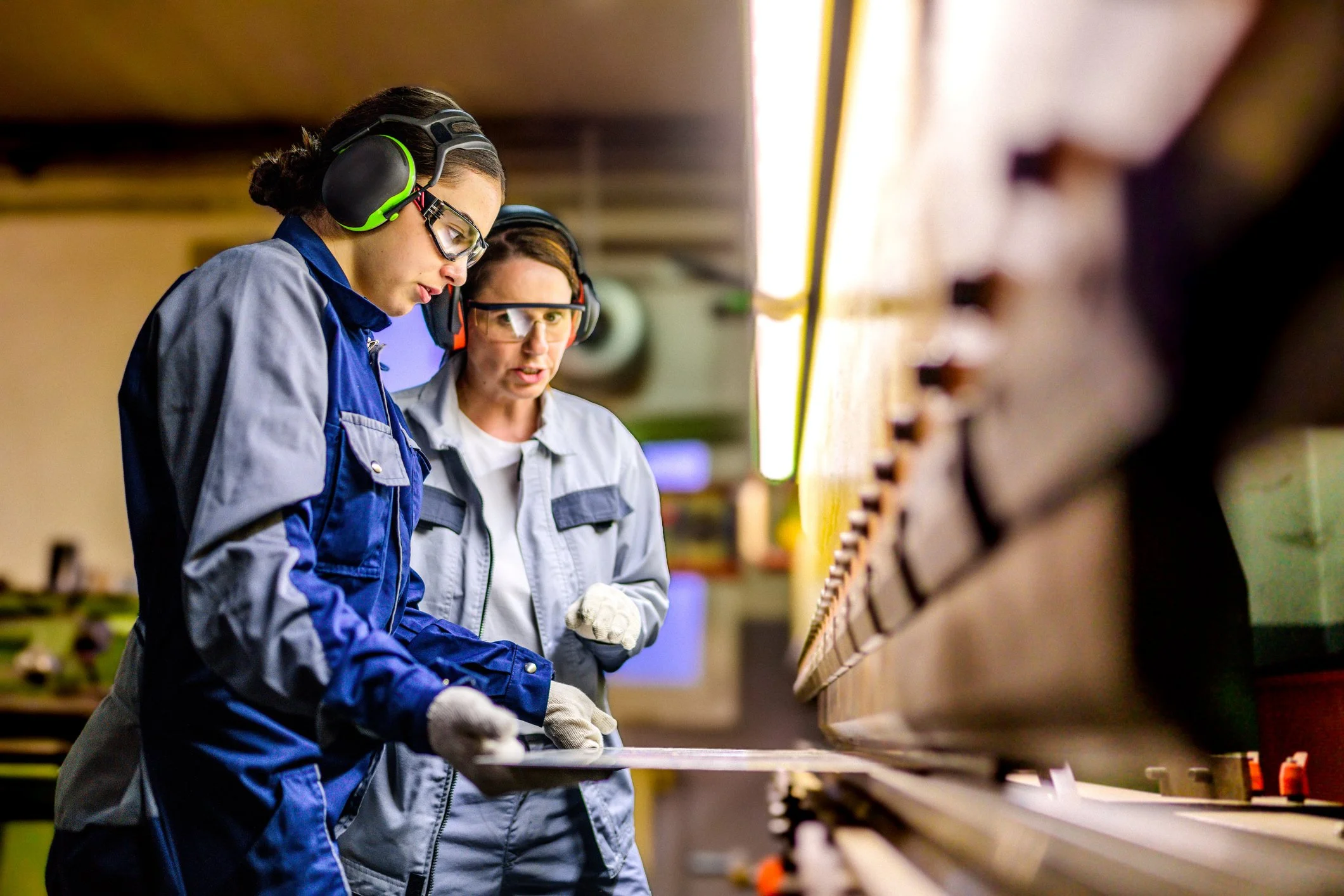 Two women working in a workshop, wearing safety glasses and ear protection, examining a piece of material.