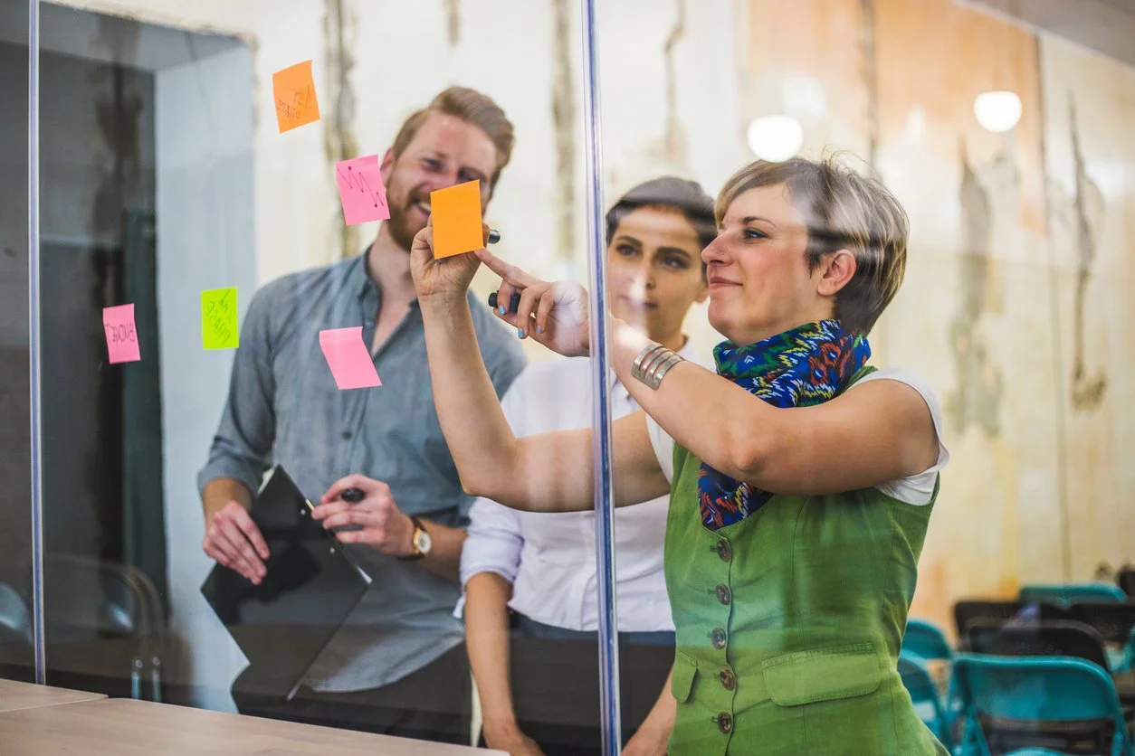 Three people working together in an office, writing on sticky notes on a glass wall, smiling and collaborating.