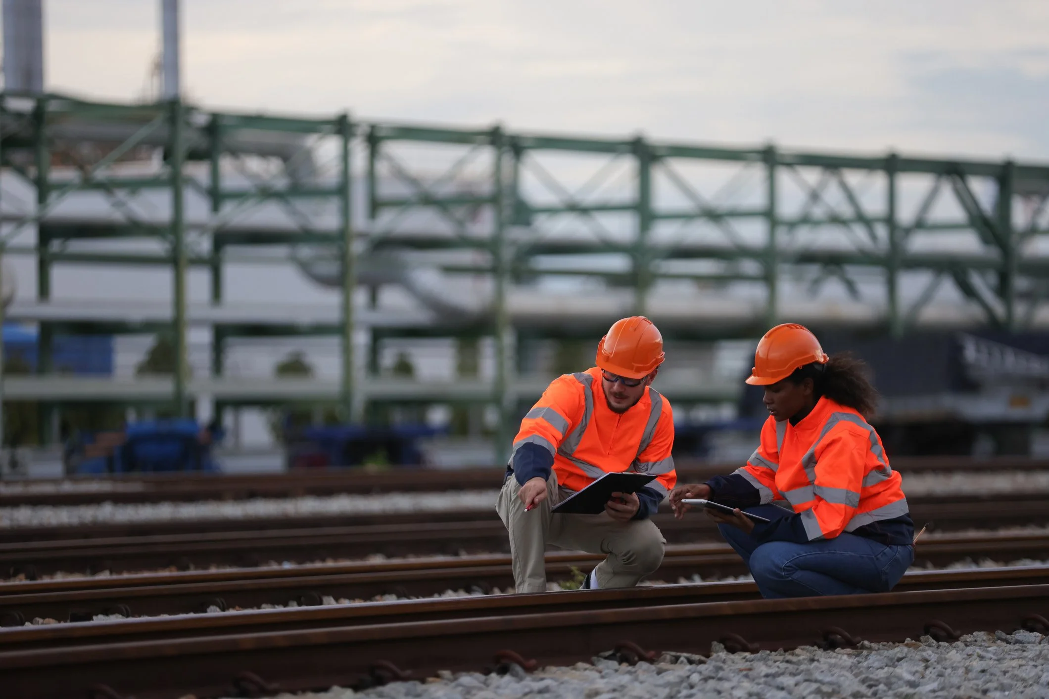 Two construction workers with orange safety vests and helmets working on railroad tracks at a construction site, with a steel scaffolding structure in the background.