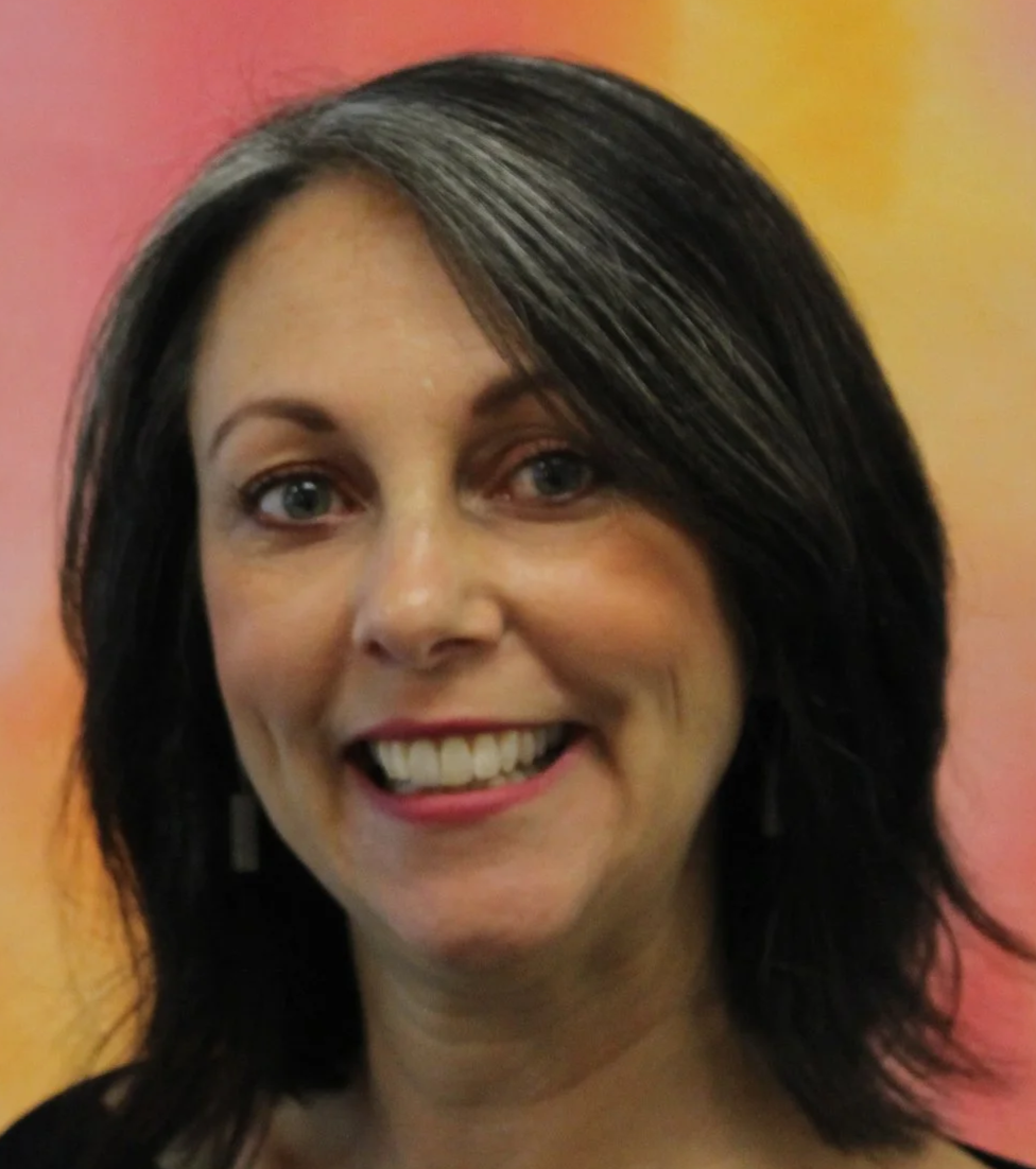 Jillian Douglas smiling with wavy, dark hair, standing in front of a colorful background.