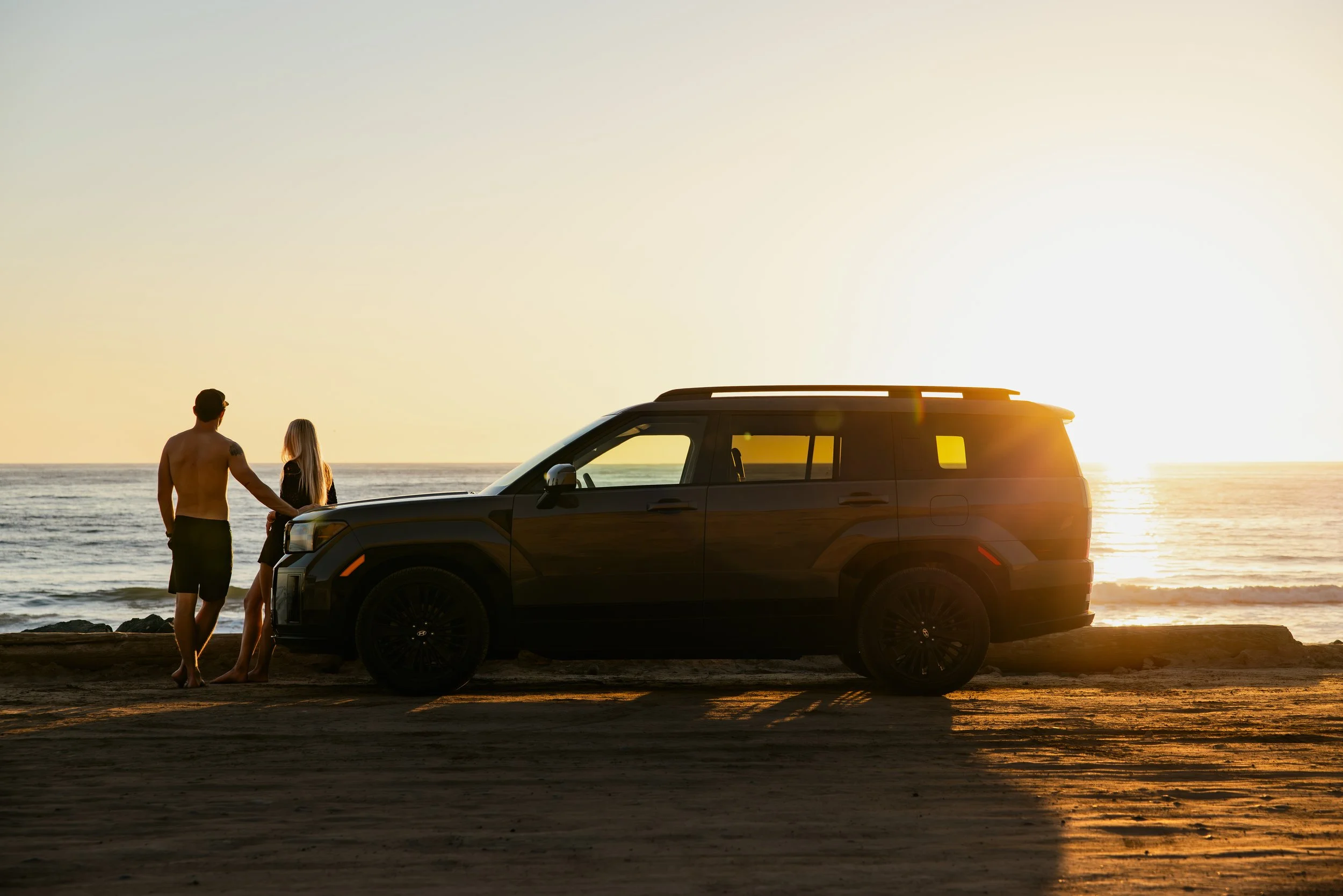A man and woman standing on a beach at sunset near a parked black SUV.