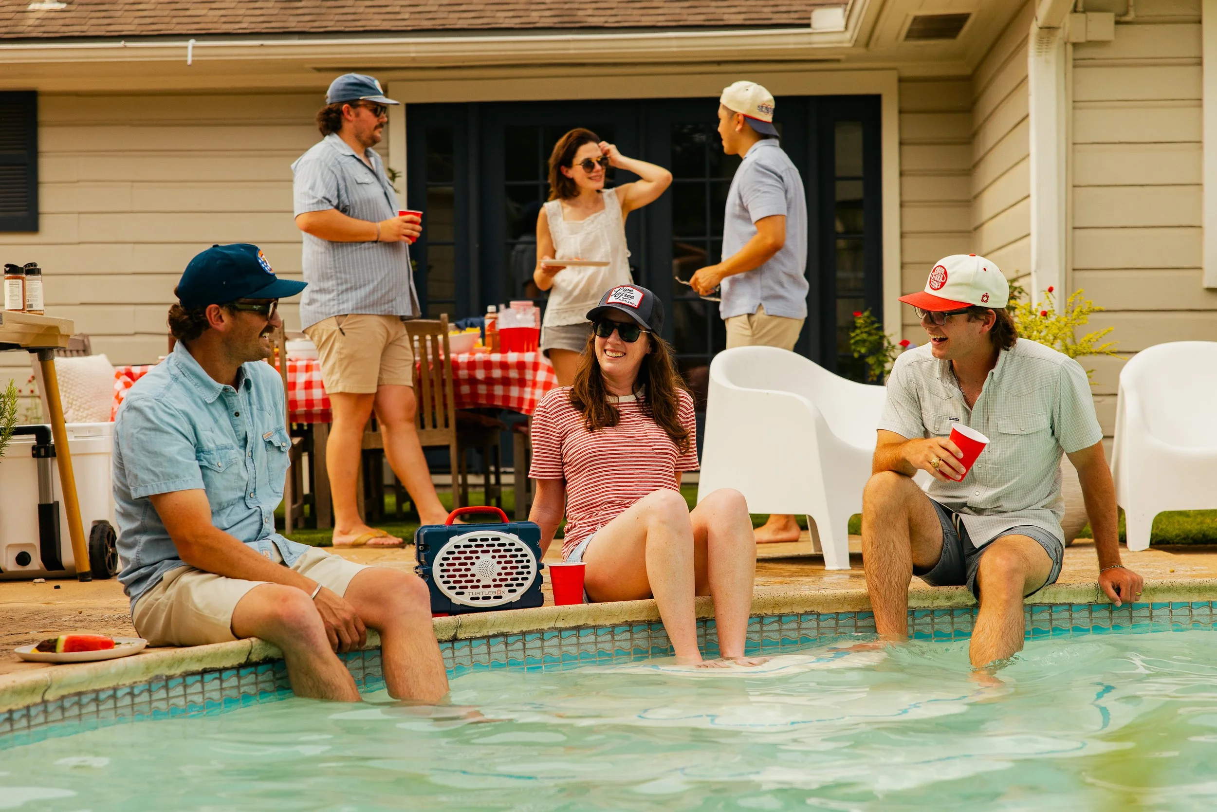 Group of friends gathered by a swimming pool, some sitting on the edge with feet in the water, others standing and talking, enjoying a sunny day with drinks and snacks.