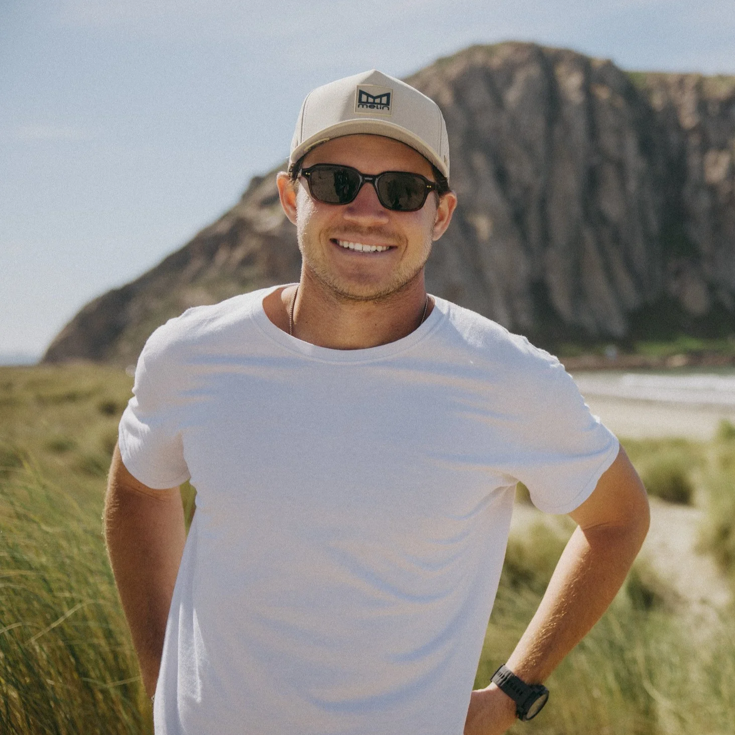 A man wearing sunglasses, a beige cap with a logo, a white t-shirt, and a watch standing outdoors with a rocky hill or mountain in the background.