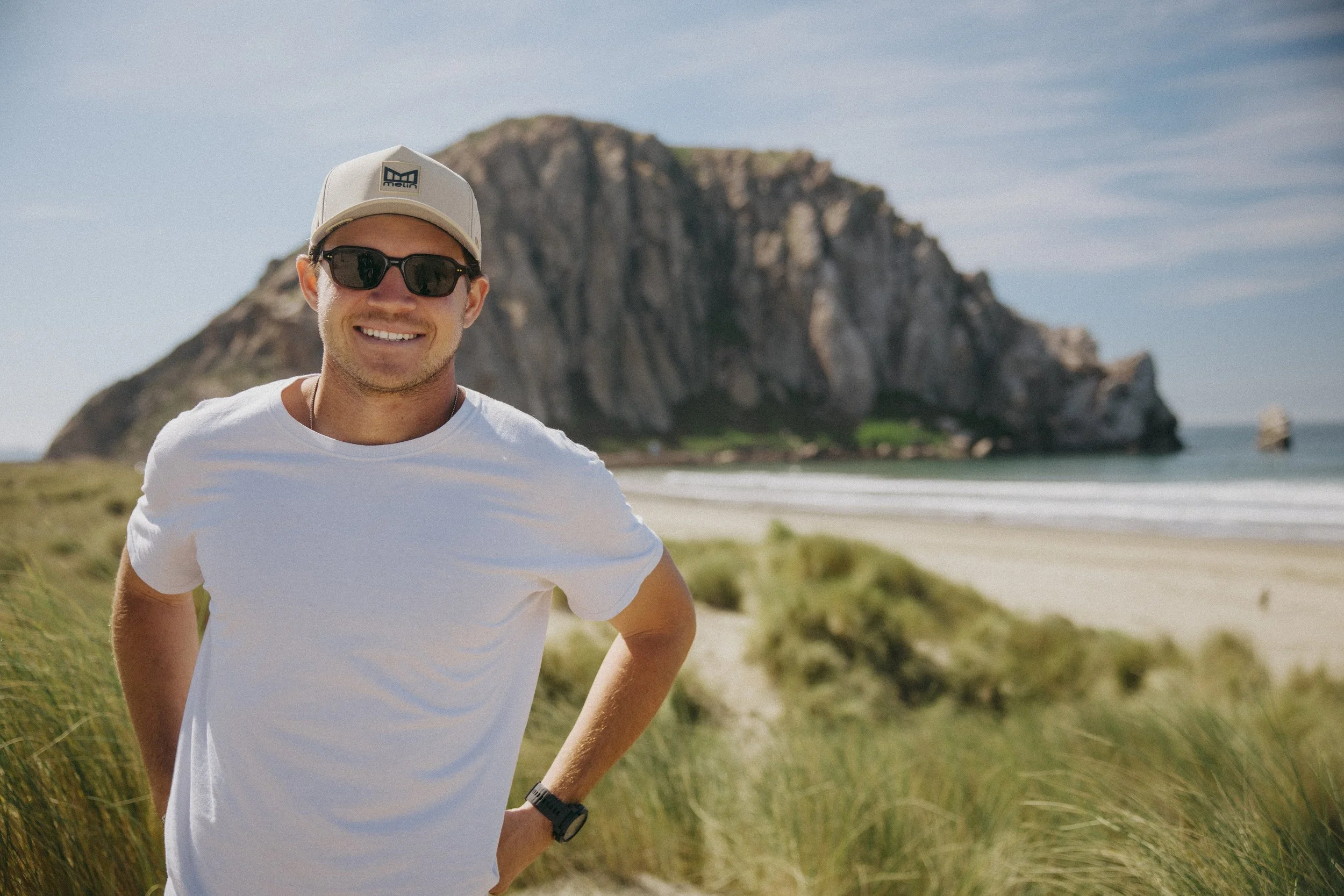 A young man smiling at the camera standing outdoors on a grassy area with a large rocky cliff in the background, wearing a white t-shirt, cap, sunglasses, and a watch.