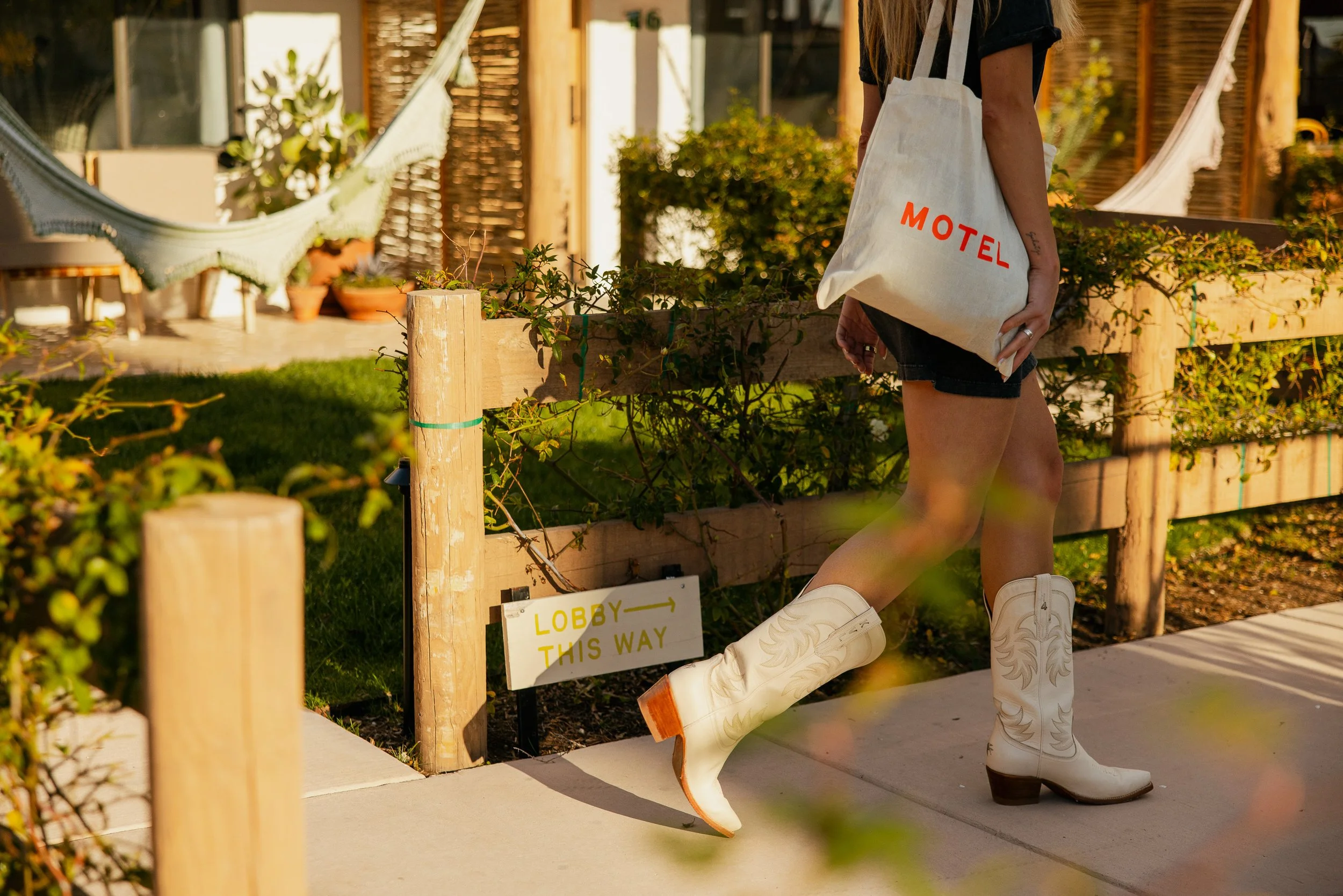 Person wearing cowboy boots and carrying a tote bag with 'MOTEL' written on it, walking past a fenced garden area with a hammock and potted plants, near a building.