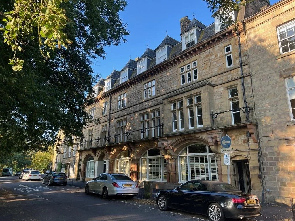 Park Parade Harrogate, street view of a multi-story stone building with bay windows and dormer windows at the top, parked cars, and green trees along the street.