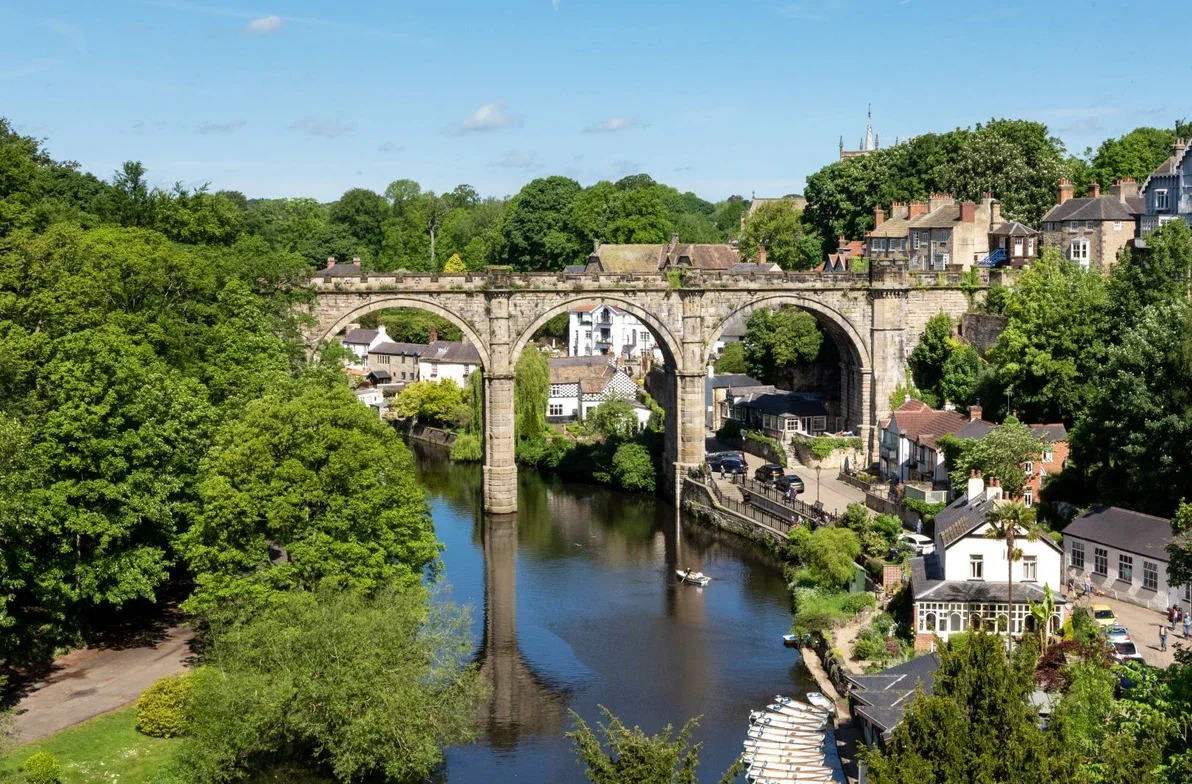 Knaresobough, A stone arch bridge crossing over a river with lush green trees on both sides. Residential houses are visible on the hillside behind the bridge.
