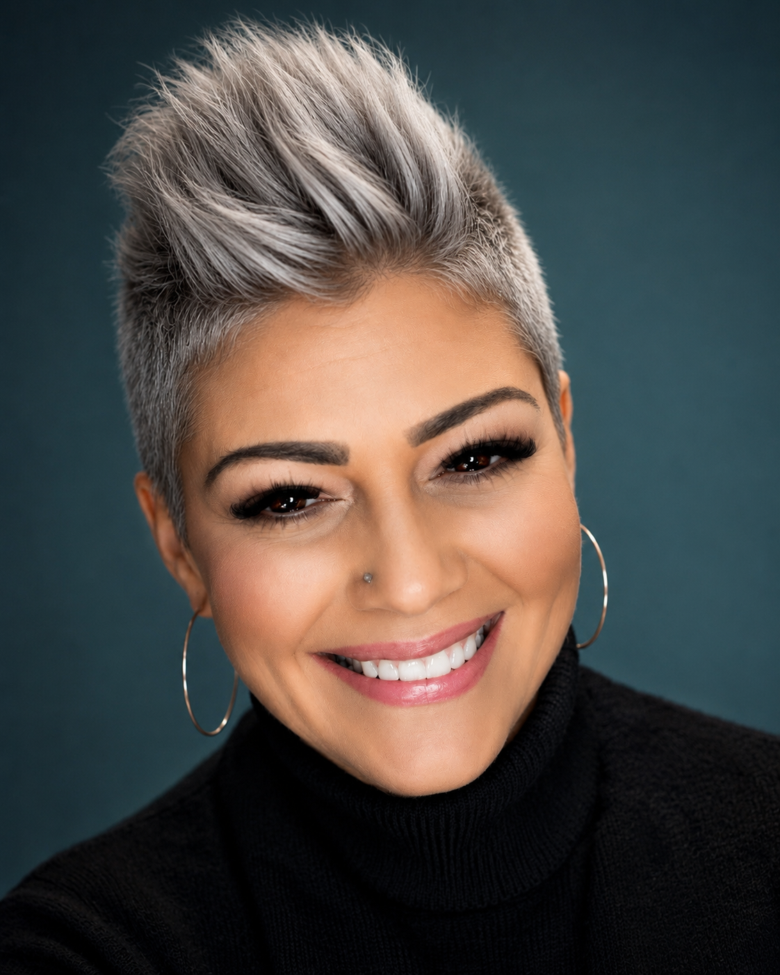 Close-up portrait of a woman with short, platinum blonde hair styled upwards, wearing a black turtleneck and hoop earrings, smiling against a dark green background.