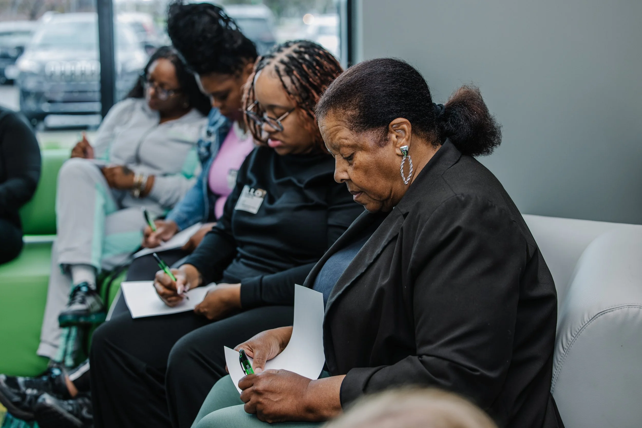A group of diverse women with different hairstyles and skin tones are sitting in a waiting area, all engaged in writing on notepads or paper.
