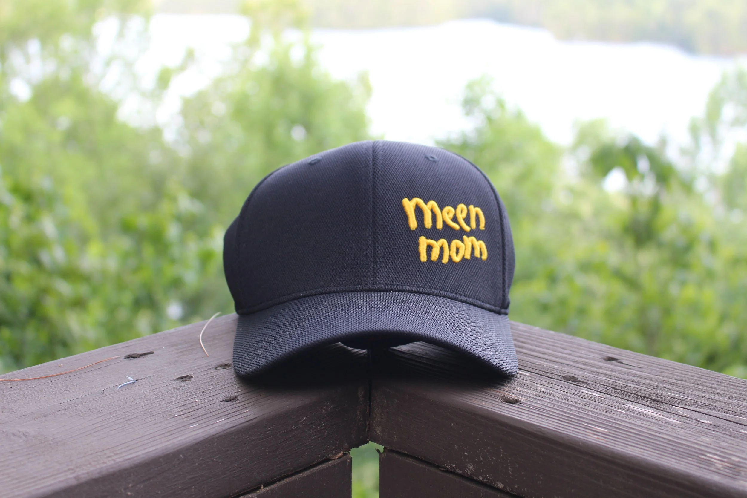 A dark blue baseball cap with yellow embroidered text that says 'Meem Mom' rests on a wooden surface outdoors, with a blurred background of green trees and sky.