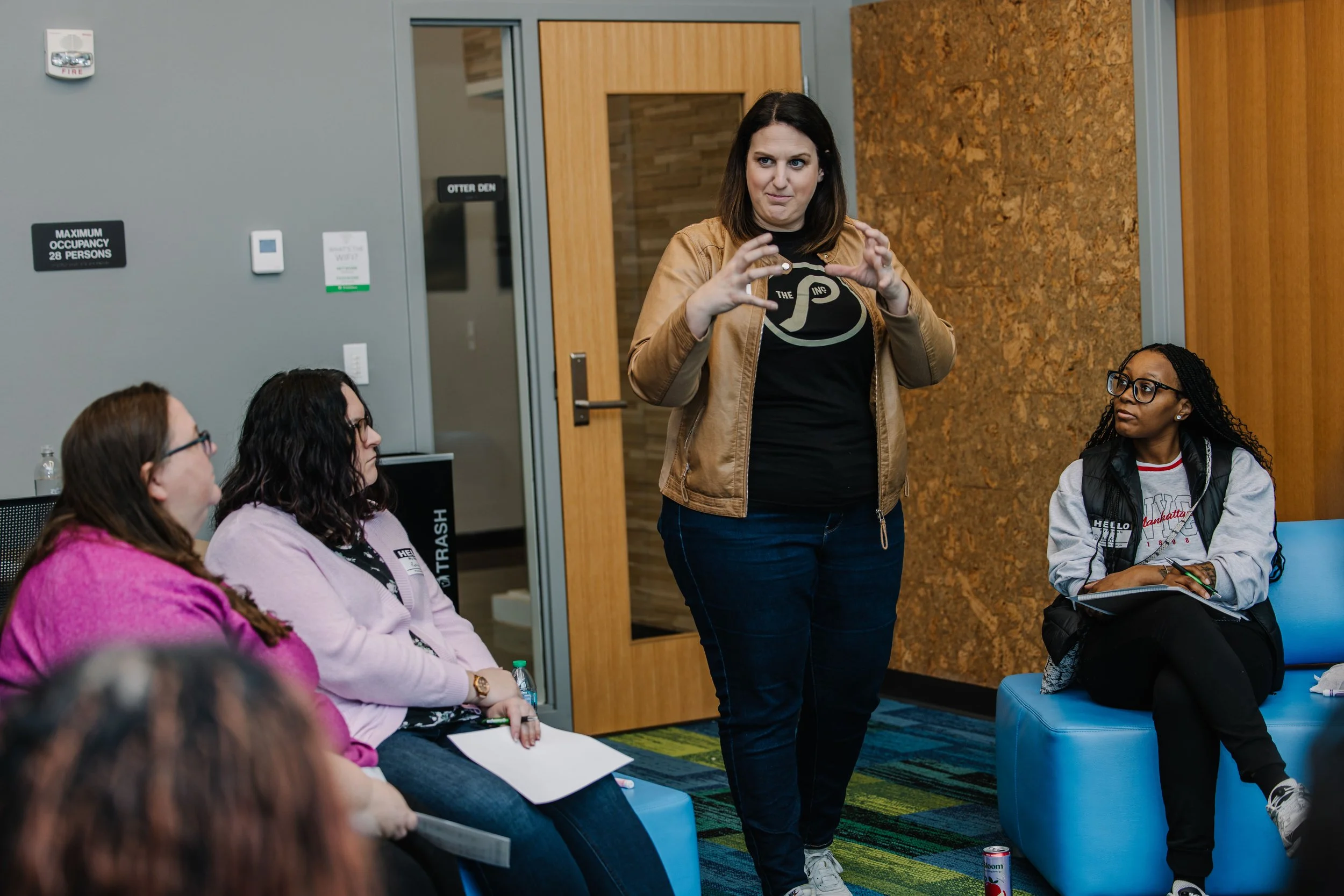 Dr. Andrea Mata standing and speaking to a group of seated women in a room with gray walls and a wooden door. The women are listening attentively, and one has a notepad and pen.