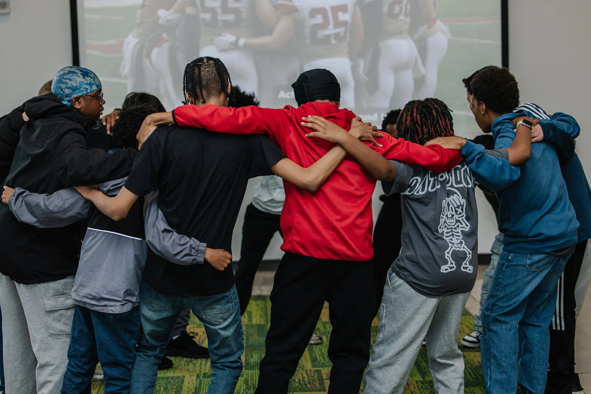 A diverse group of young people in sports attire standing in a huddle with their arms around each other, watching a football game on a screen.