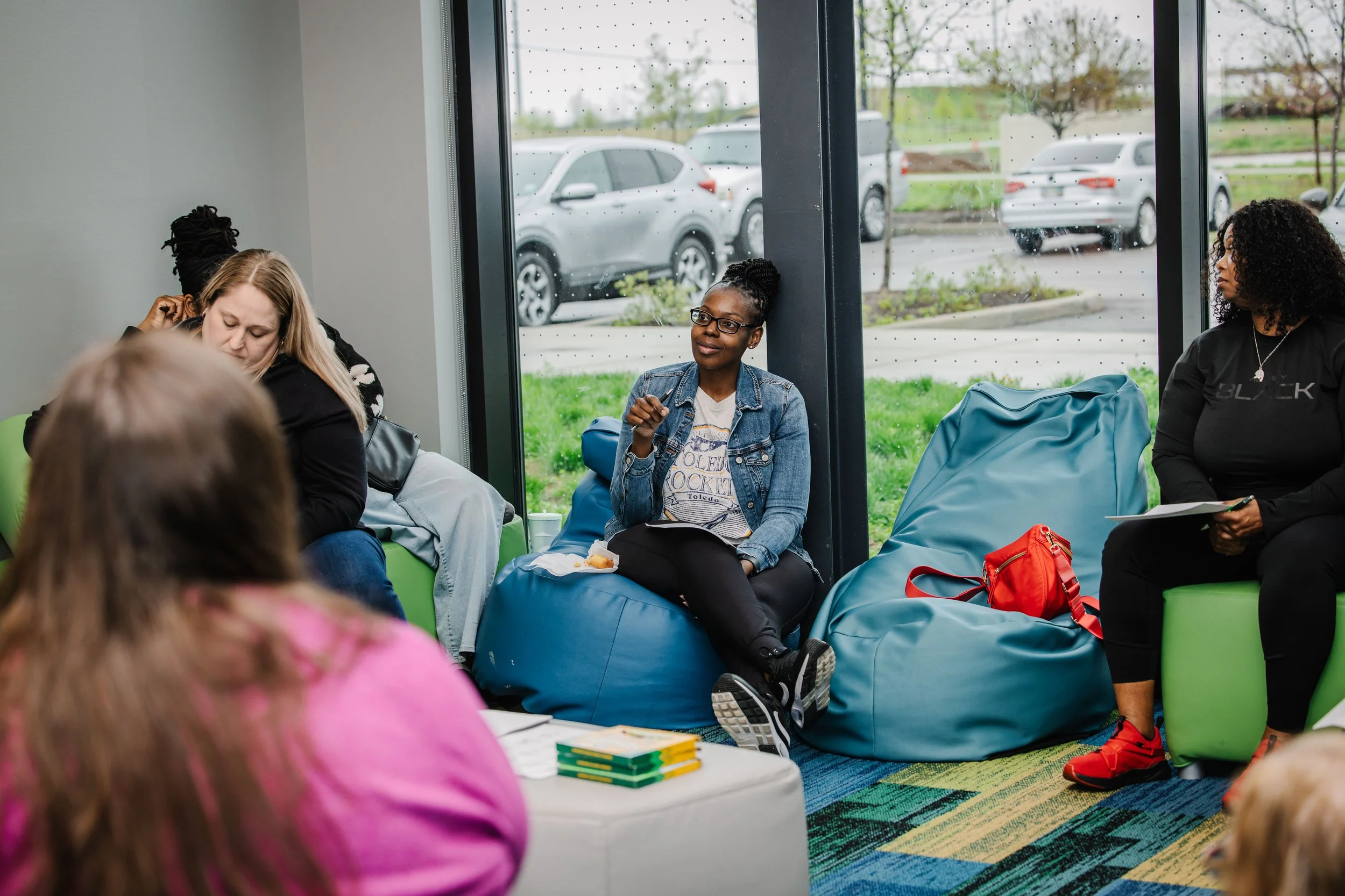 Group of diverse women sitting and talking in a modern indoor space with large windows and cars outside.