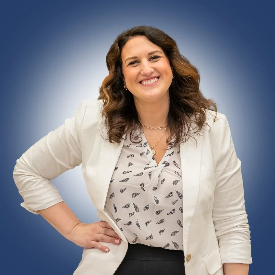 Dr. Andrea Mata smiling and posing with one hand on her hip, wearing a white blazer, patterned blouse, and black pants, against a blue gradient background.