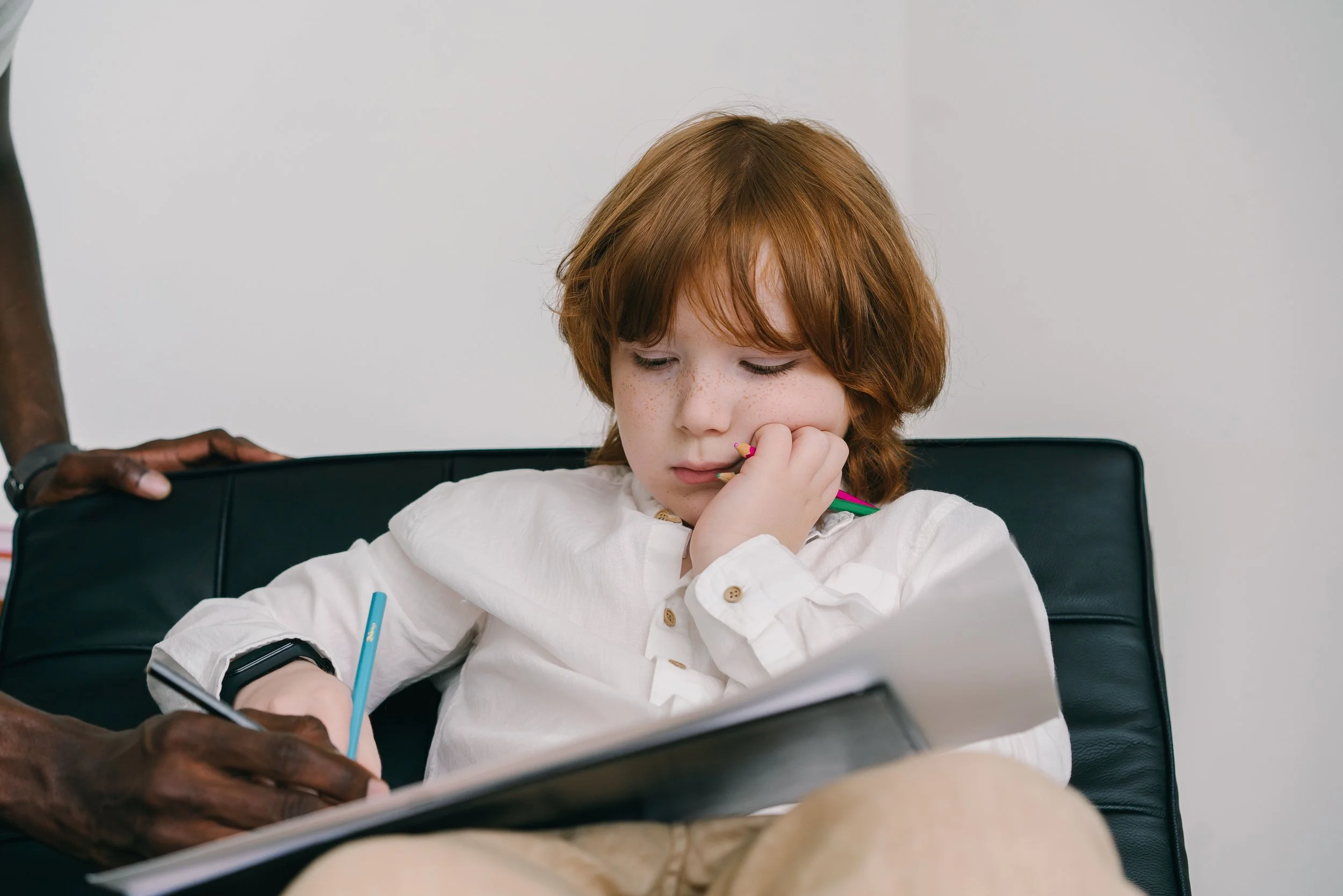 A young red-haired girl with freckles, sitting at a table with a tablet computer, holding a pink-colored pencil, appearing focused or thoughtful.