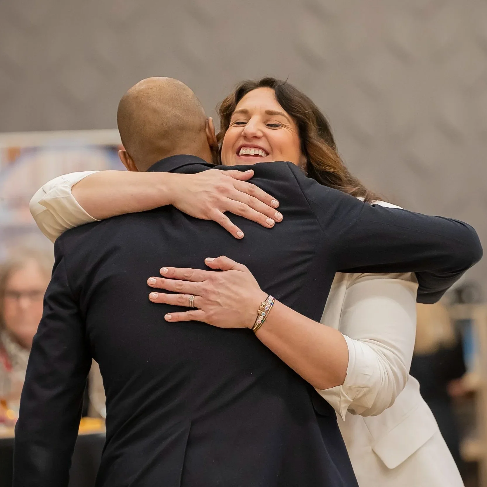 Dr. Andrea Mata smiling while hugging a man in a black suit, with her arms around his shoulders, in an indoor setting.