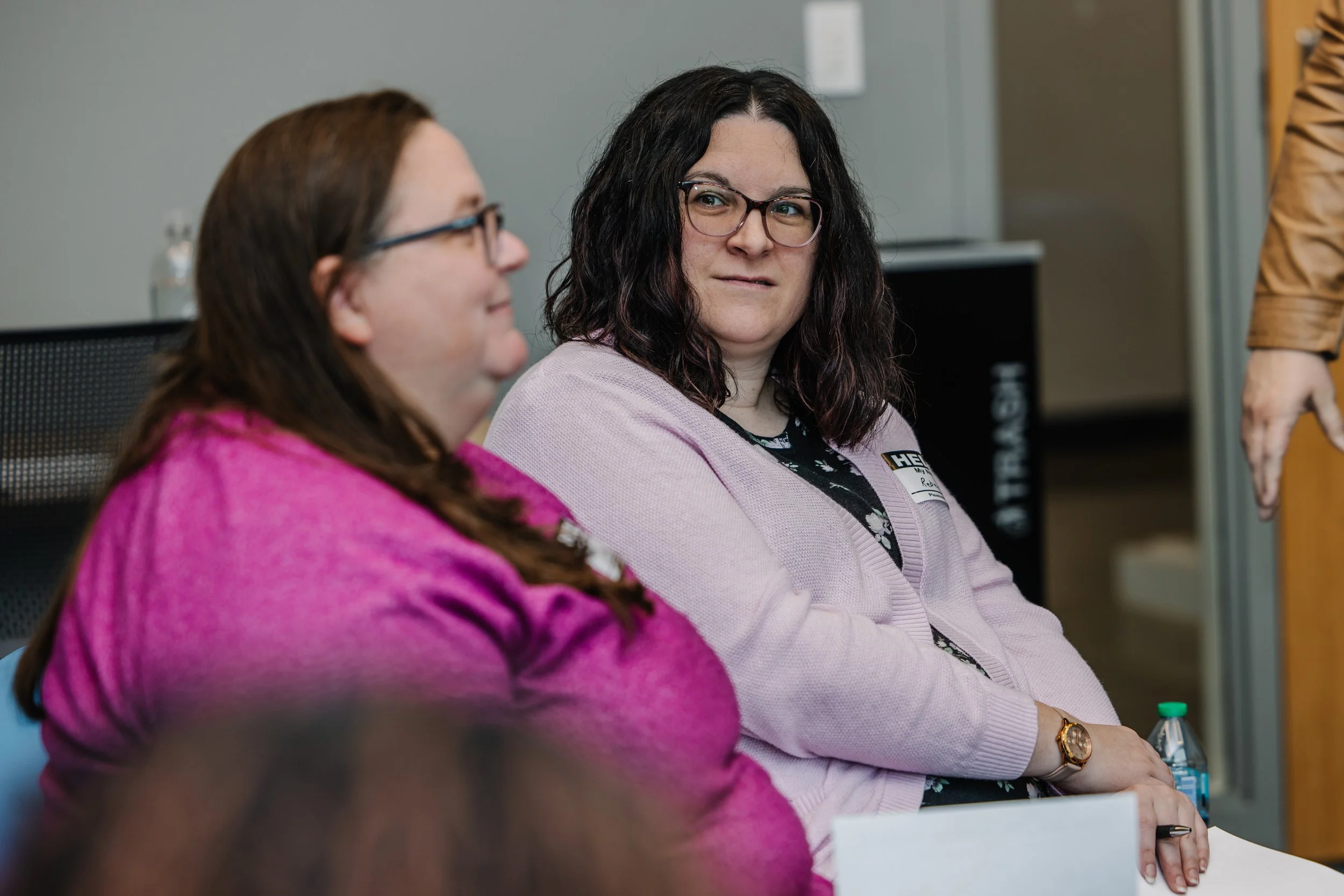 Two women sitting in a meeting room, engaged in conversation. One woman has dark, curly hair and glasses, wearing a pink cardigan. The other woman has long, straight hair, glasses, and wears a purple top.