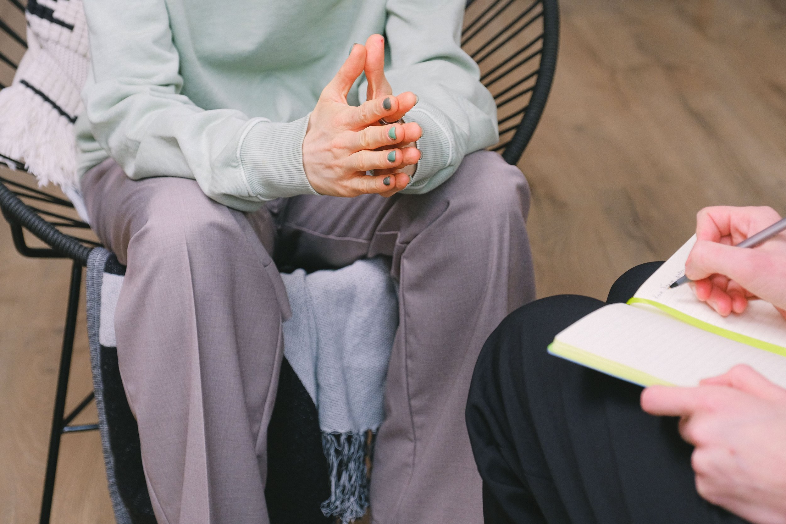 Person sitting in a wheelchair with hands in a prayer position, wearing a light green sweatshirt and gray pants, during a counseling session with a notepad and pen.