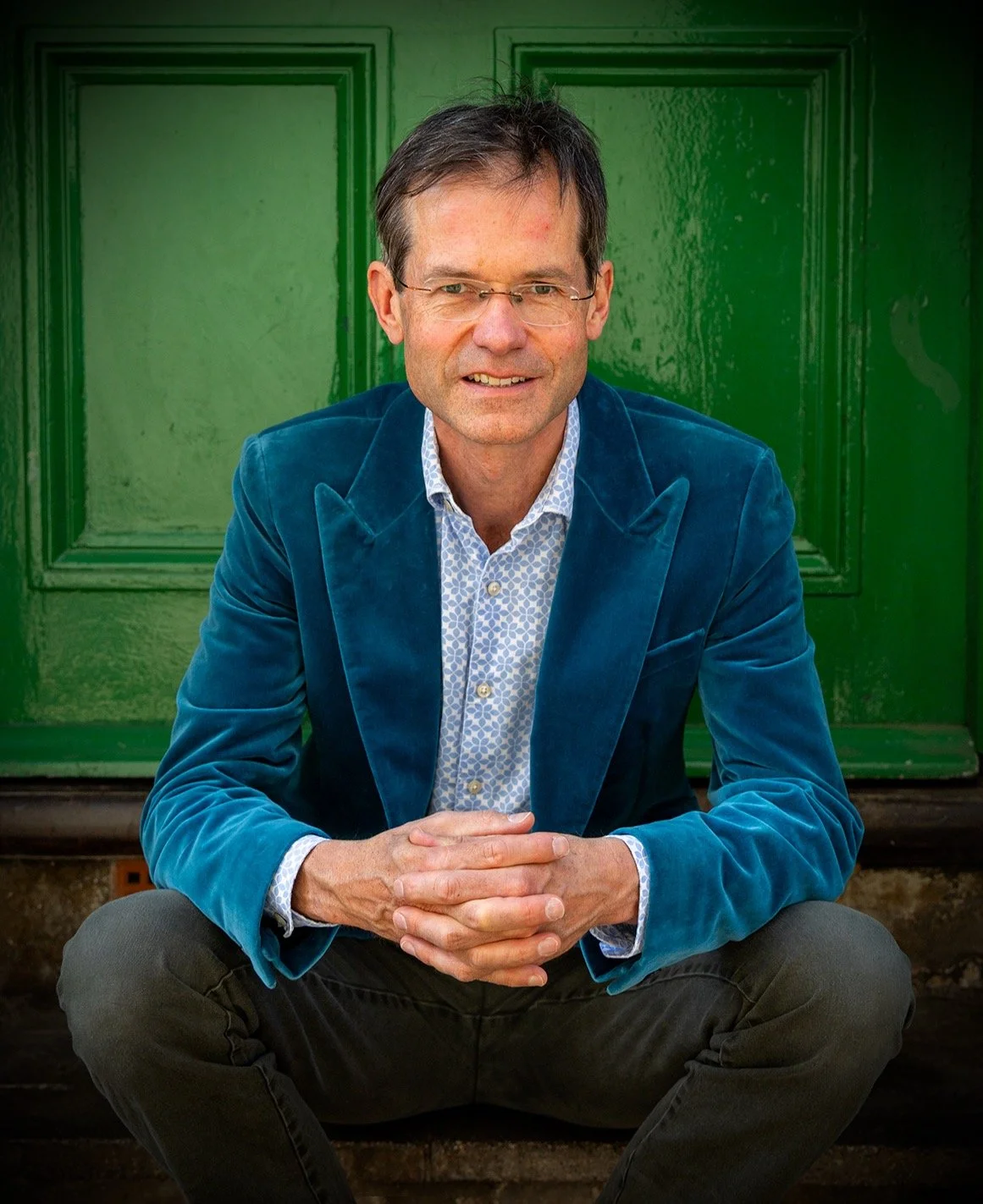 A man with glasses, wearing a blue velvet blazer and a patterned shirt, sitting on steps in front of a vibrant green door.