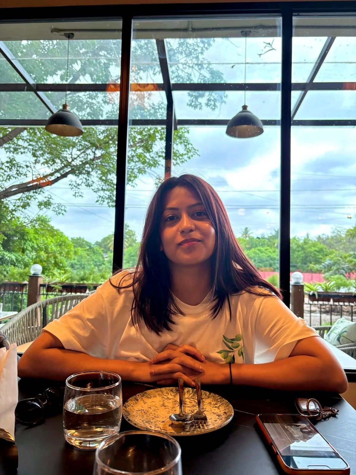 Young woman with dark hair sitting at a restaurant table with a plate, drinking glass, and phone, inside a glass-enclosed space with trees and cloudy sky visible outside.