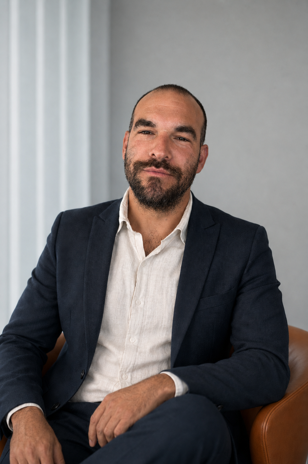 A man with a beard and short hair, dressed in a navy blazer and white shirt, sitting on a chair against a plain background.