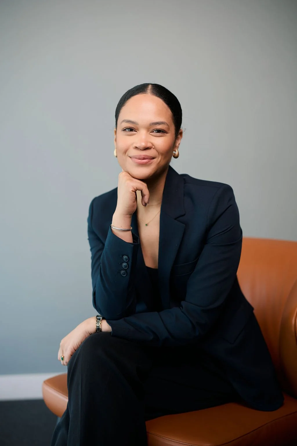 Woman in a navy blue suit sitting on a leather chair, smiling with hand on chin.