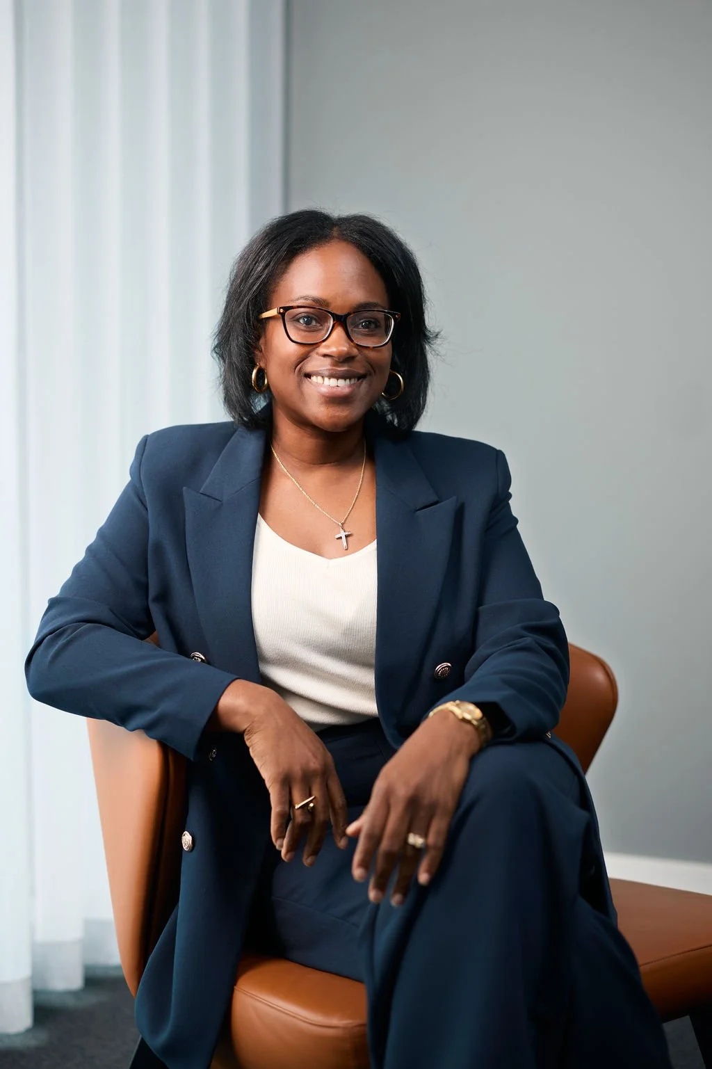 Person wearing a dark blue suit sitting on a brown chair, smiling, with short black hair and glasses.