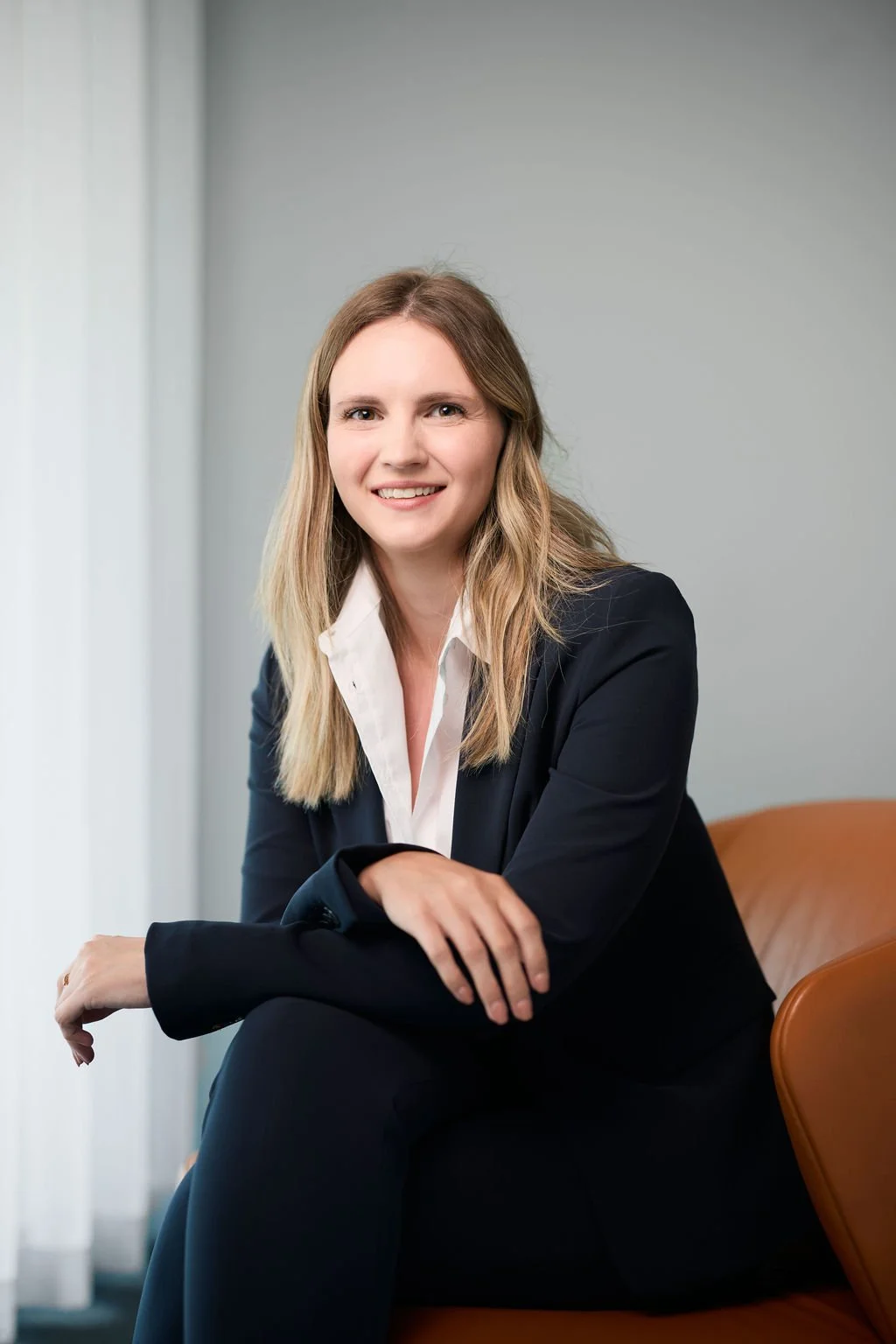 A woman with blonde hair, wearing a black suit and white shirt, is sitting on a brown chair. She is smiling and has one arm resting on her knee.