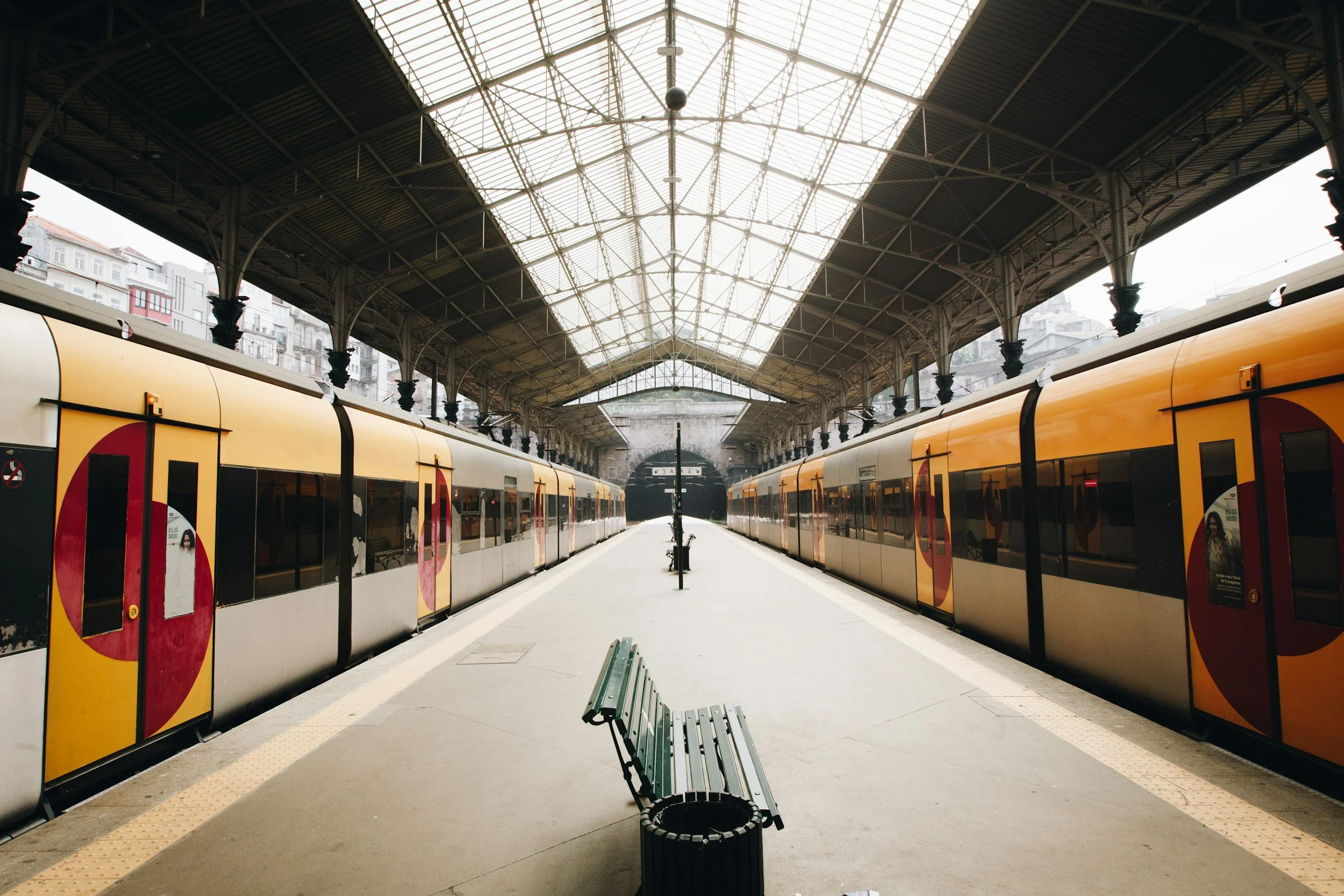 Empty train station platform with yellow trains on either side, a metal bench in the center, under a large glass and metal roof, with buildings visible in the background.