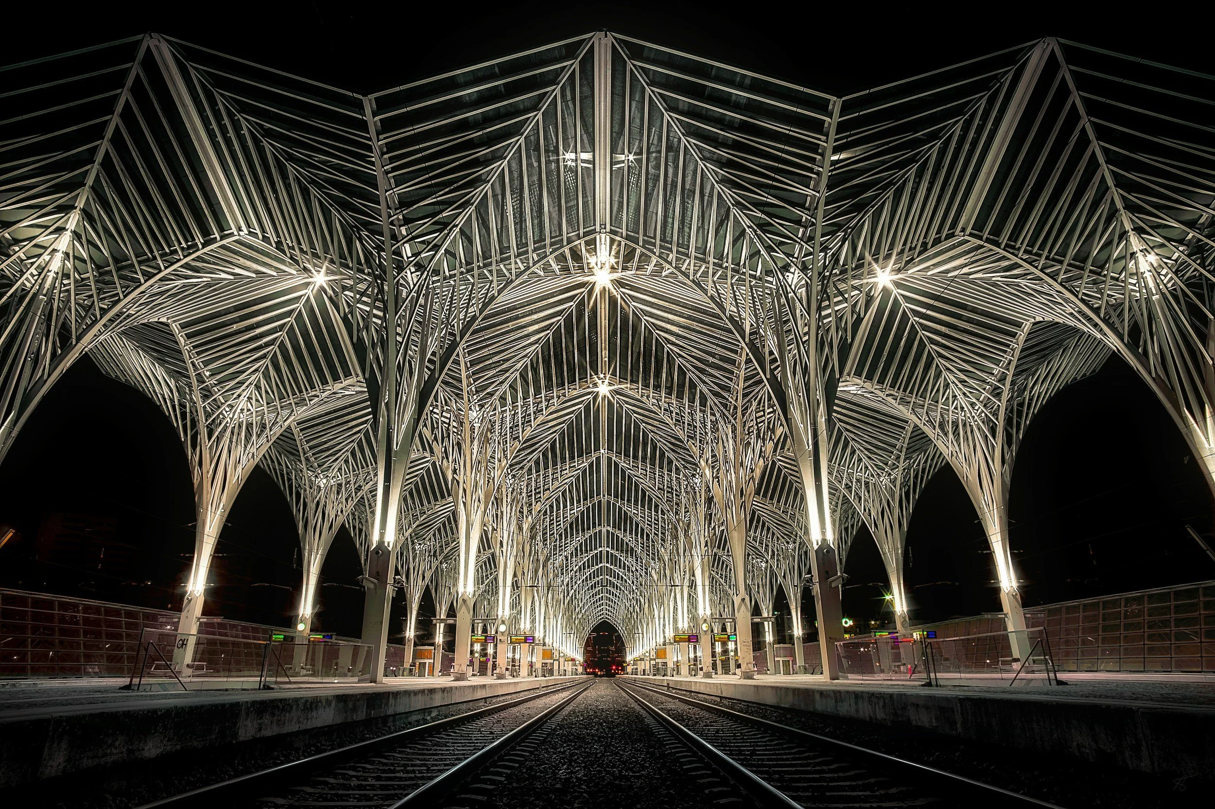 Night view of a modern metal architectural structure at a train station, illuminated with bright lights, with train tracks in the foreground.