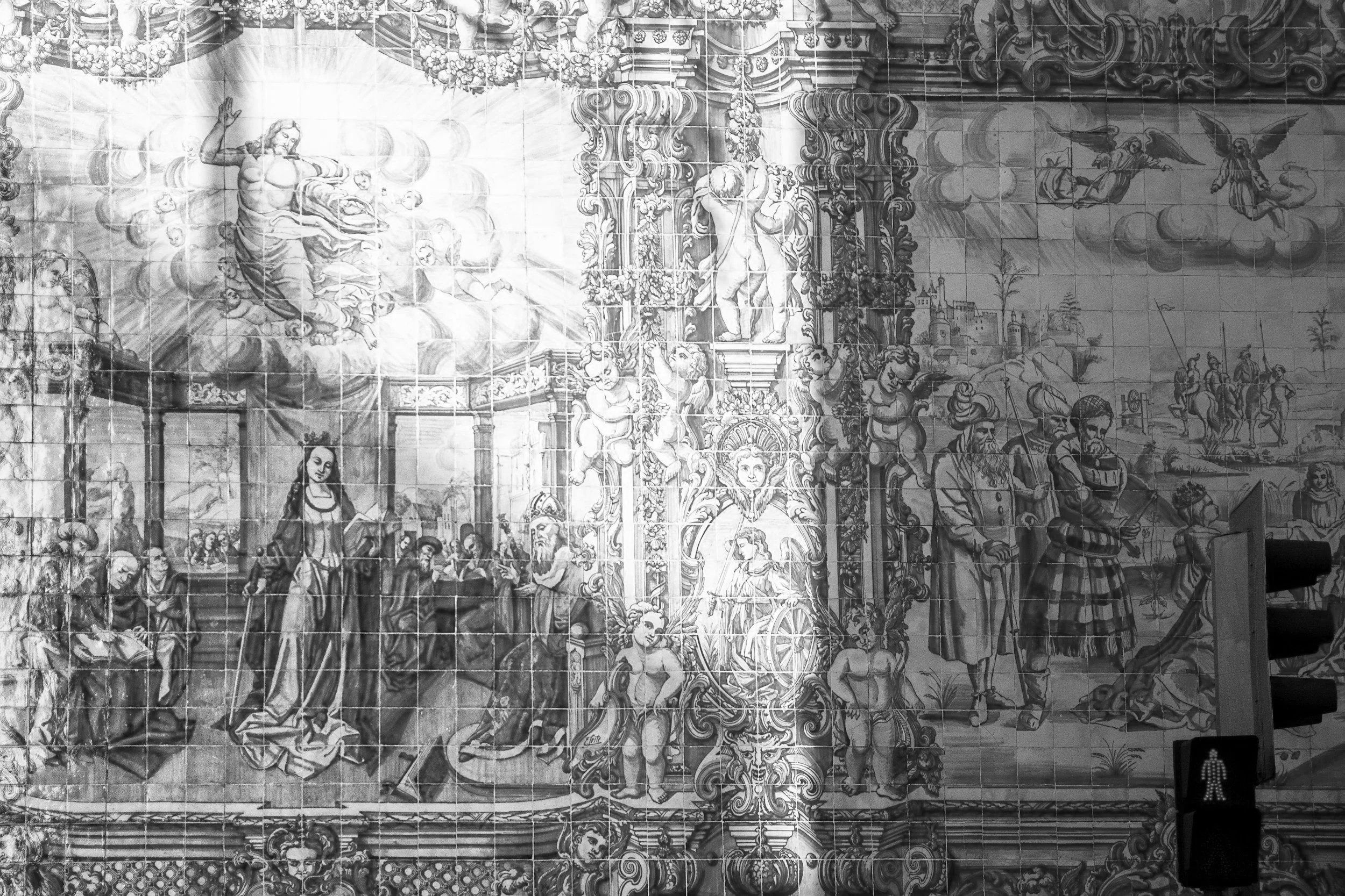 A large decorative tile mural depicting a classical scene with angelic figures, a woman playing a musical instrument, and a landscape with buildings and trees in the background.