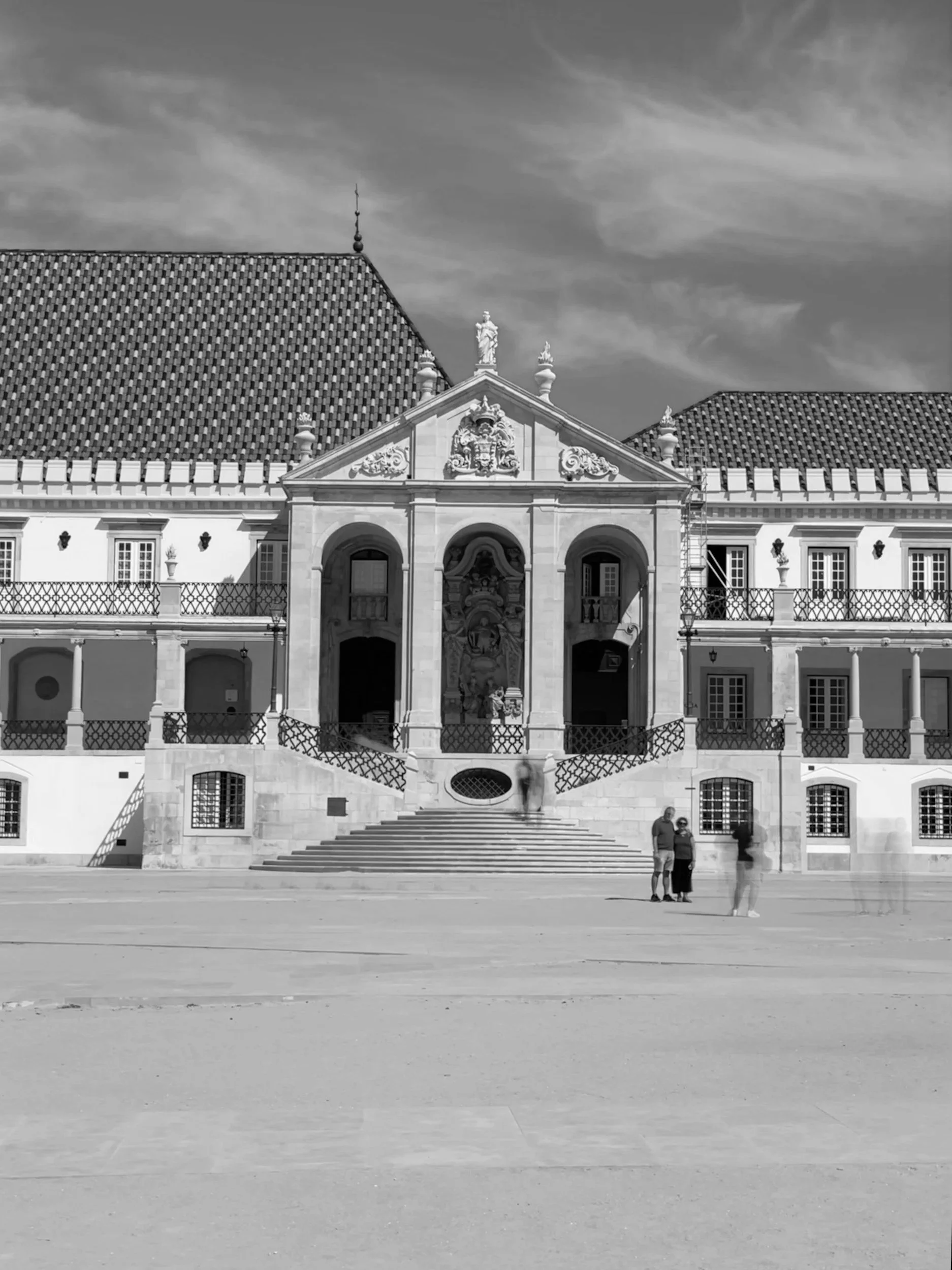 A black and white photo of a historic building with ornate architectural details, steps leading up to the entrance, and a large open courtyard in the foreground. Three people are standing at the bottom of the stairs.
