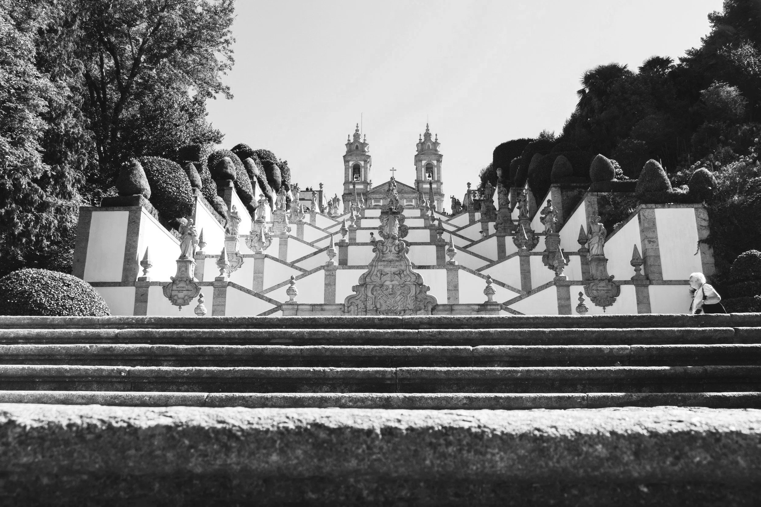 Black and white photo of an ornate staircase with multiple statues along the sides, leading up to a church with twin bell towers and a cross at the top, surrounded by trees.