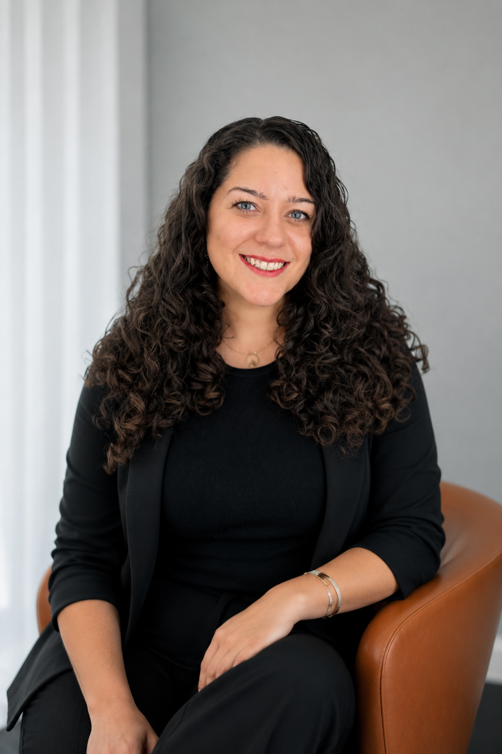 Portrait of a woman with long curly dark hair, wearing a black blazer and black top, smiling while sitting on a tan chair in a room with light-colored walls and white curtains.