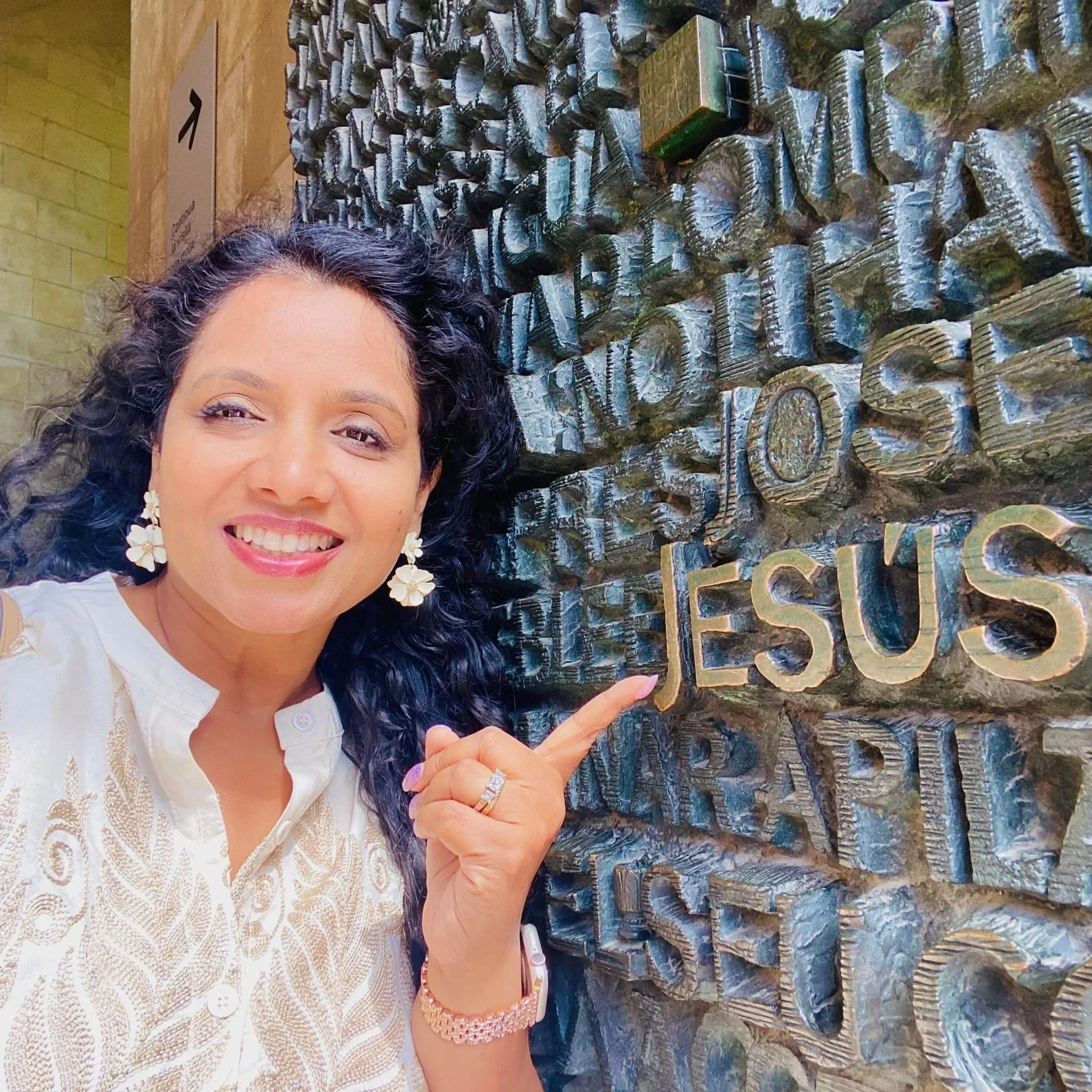 Close-up of a woman smiling, wearing flower earrings and jewelry, pointing at a textured stone wall with Jesus carved lettering.