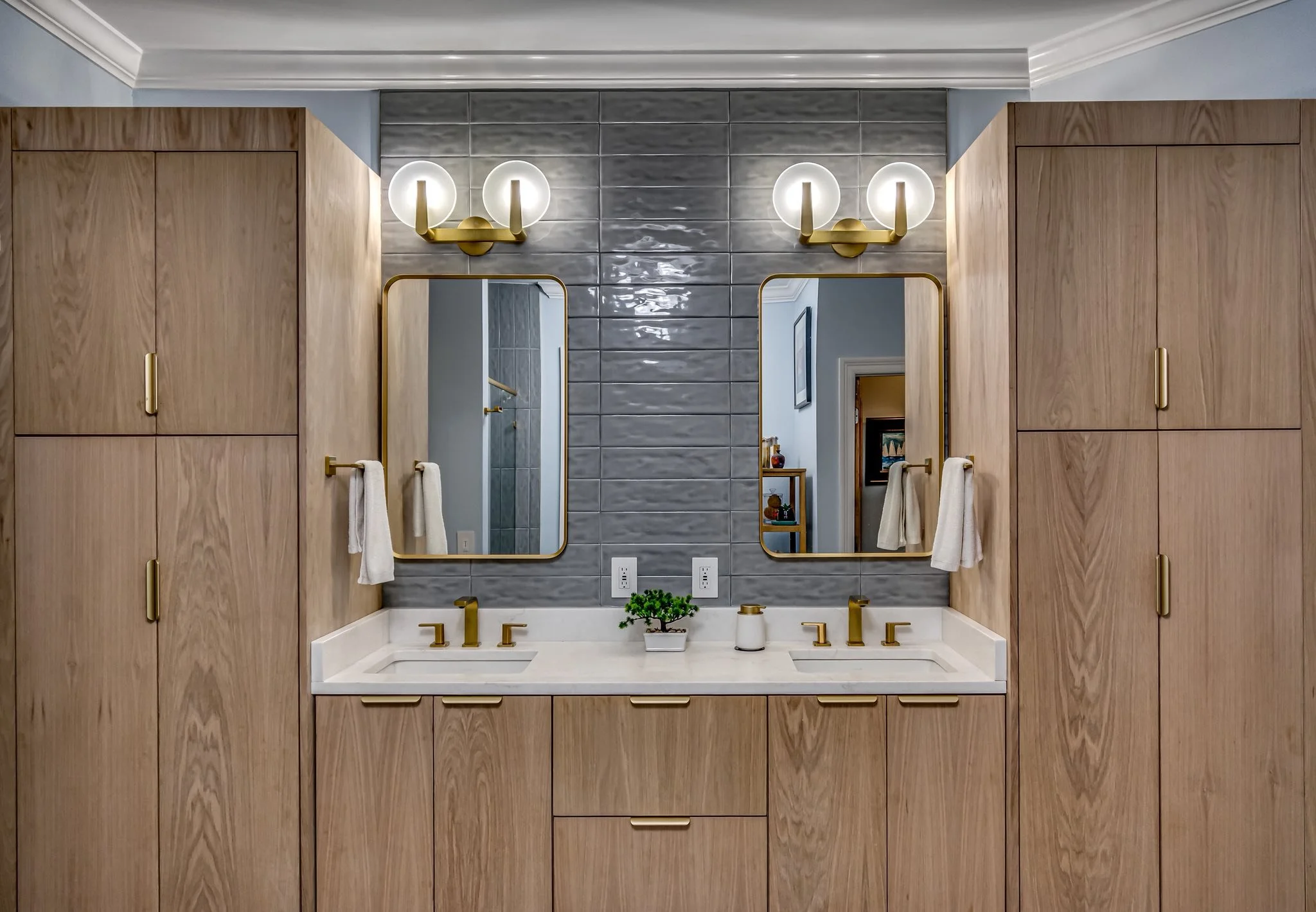 A modern bathroom vanity with a light wood cabinet, gold fixtures, two mirrors, and two wall-mounted light fixtures, with a small potted plant and soap dispenser on the white countertop.