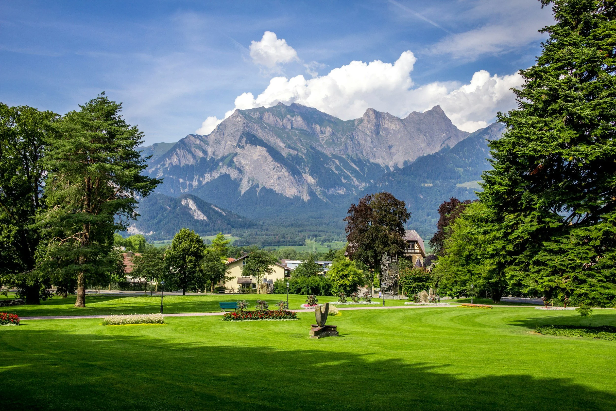A scenic park with lush green grass, colorful flower beds, and tall trees, with mountains in the background and a partly cloudy sky.