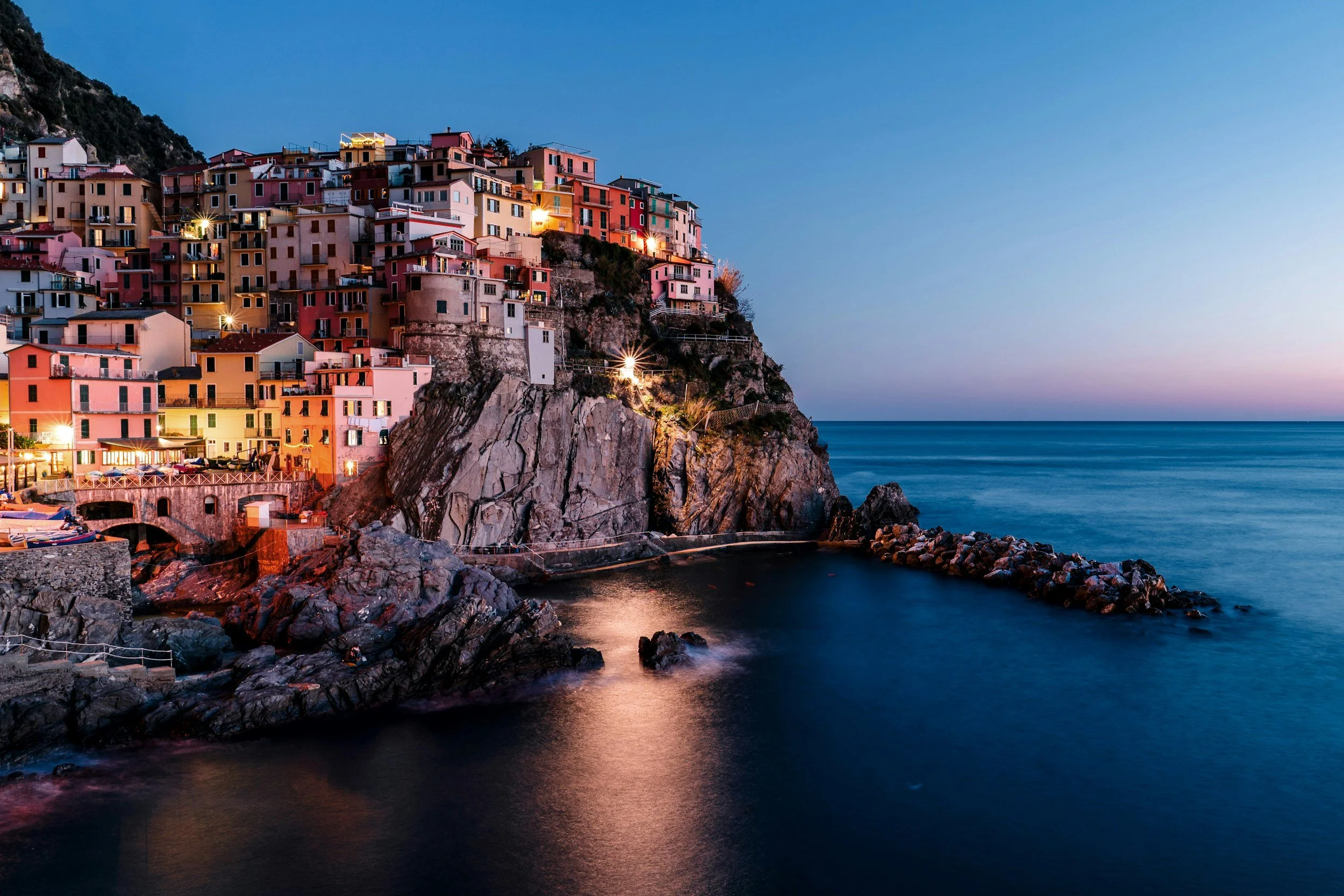 Colorful houses built on a hillside overlooking the ocean during twilight, with lights illuminating the buildings and calm water reflecting the scene.