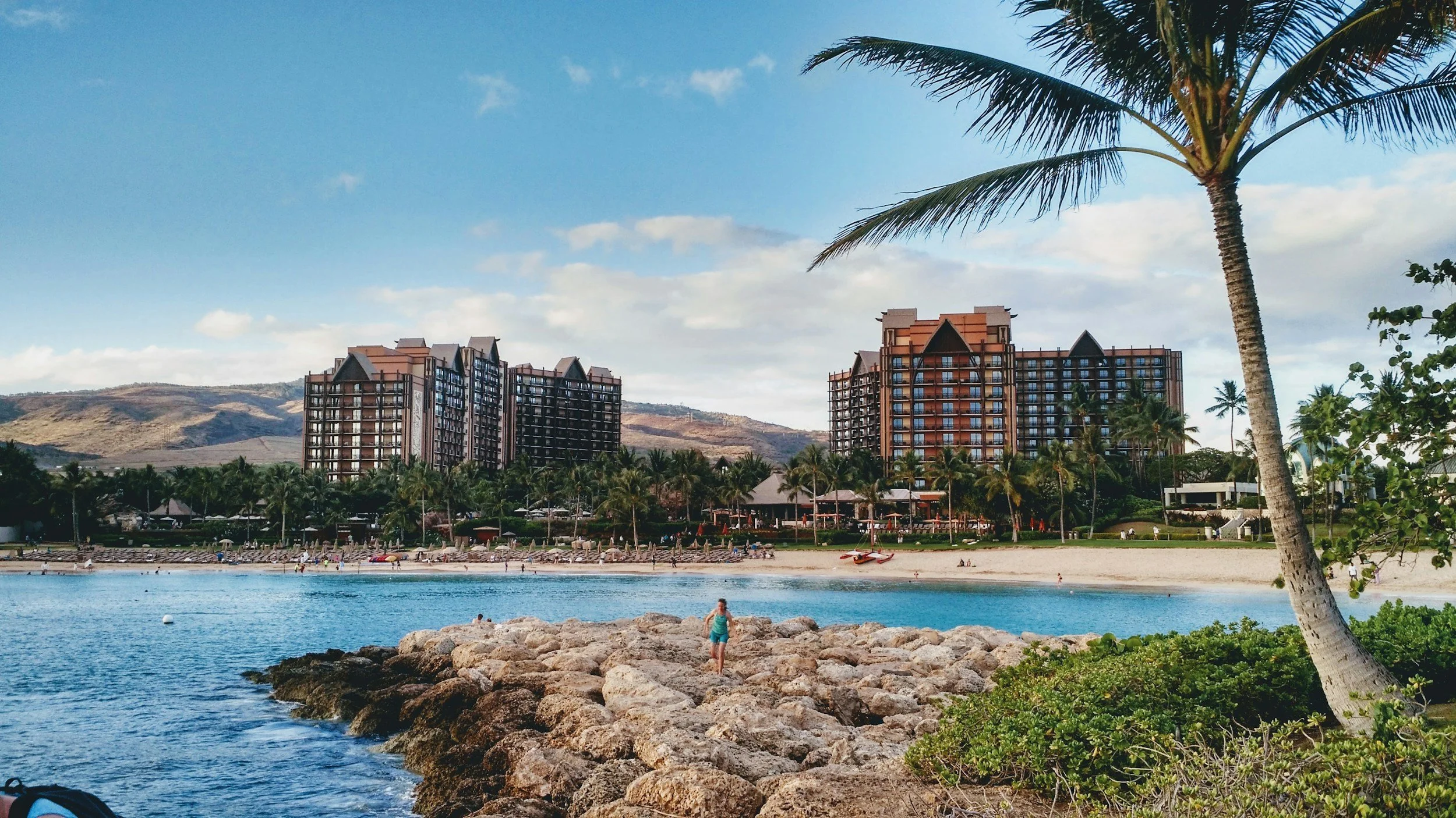 A beach scene with a person standing on rocks near the water, palm trees, and high-rise buildings in the background under a partly cloudy sky.